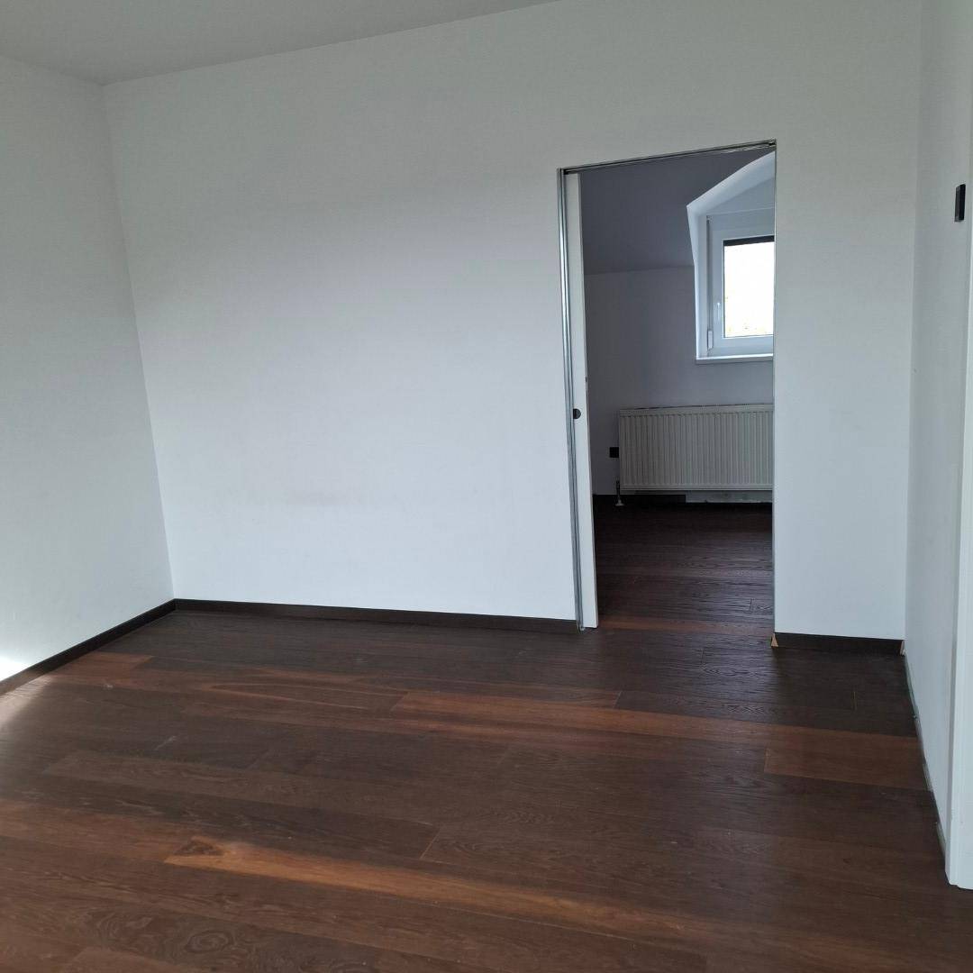 Schlafzimmer mit dunklem Holzboden und weißen Wänden, mit Zugang zu einem weiteren Raum mit Fenster.