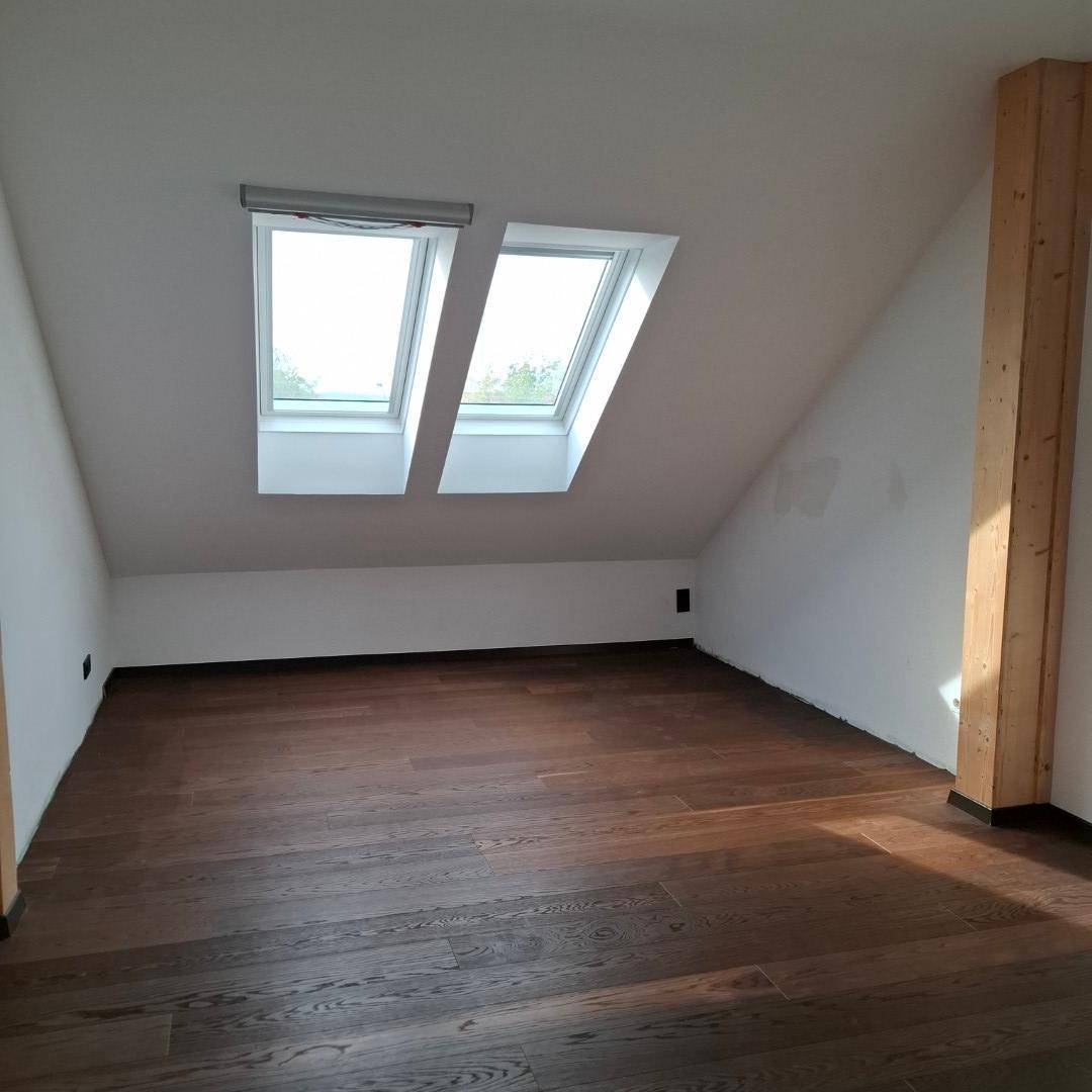Schlafzimmer mit dunklem Holzboden, weißen Wänden und zwei Dachfenstern, mit sichtbarem Holzbalken.