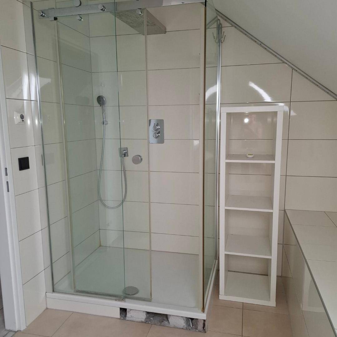 Badezimmer mit geräumiger Glasduschkabine und kleinem Regal, hell gefliest unter einer Dachschräge.
