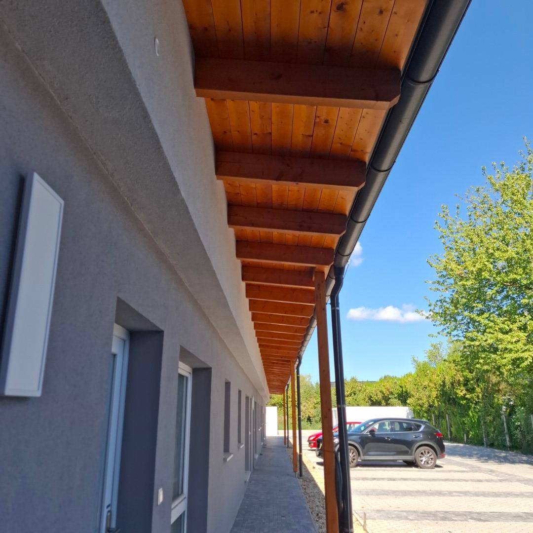 Überdachter Gang mit Holzbalken und einer Regenrinne, der zu Parkplätzen führt.