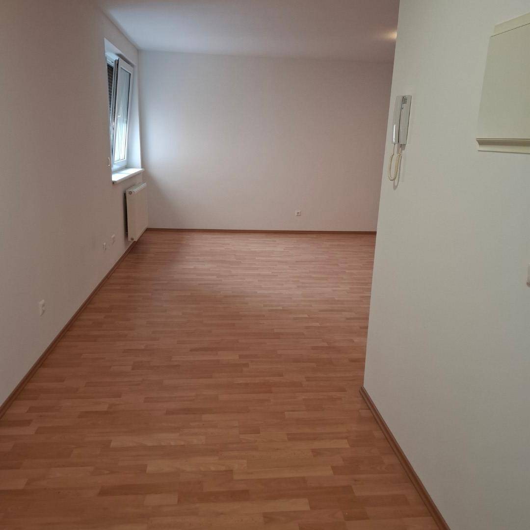 Leerer Wohnraum mit hellem Laminatboden, weißen Wänden und einem Fenster mit Heizkörper.