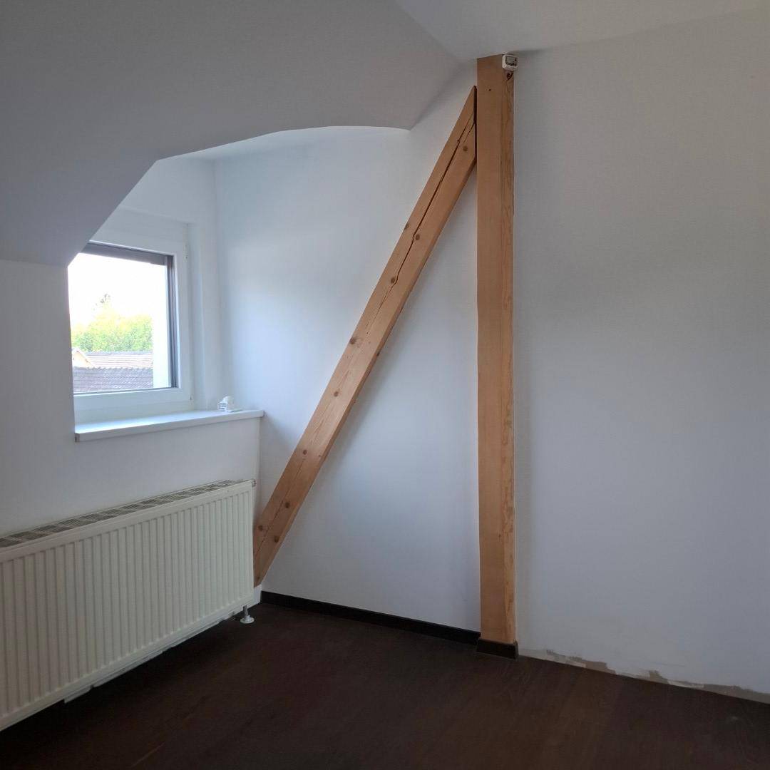 Helles Zimmer mit Holzbalken, Fenster, Heizkörper und dunklem Holzboden.