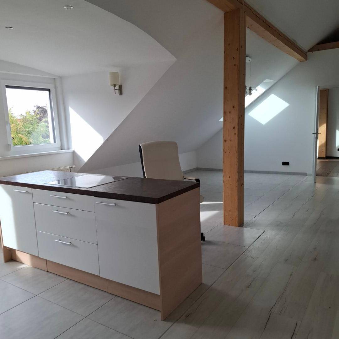 Offener Wohnbereich mit kleiner Küchenzeile, hellem Laminatboden und sichtbarem Holzbalken unter Dachschrägen.