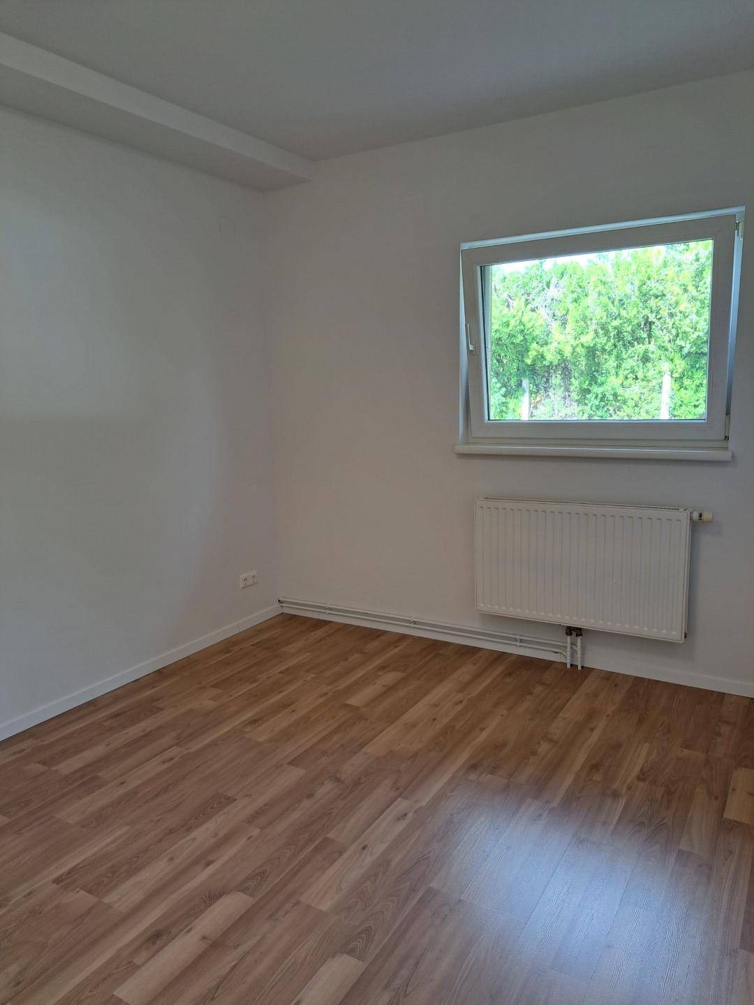Leeres Schlafzimmer mit Laminatboden, einem Fenster und einem Heizkörper unter dem Fenster.