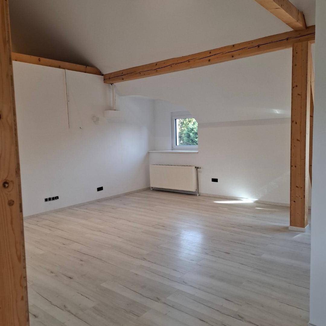 Geräumiges Zimmer mit Holzbalken, Dachschräge, Fenster und hellem Holzboden.
