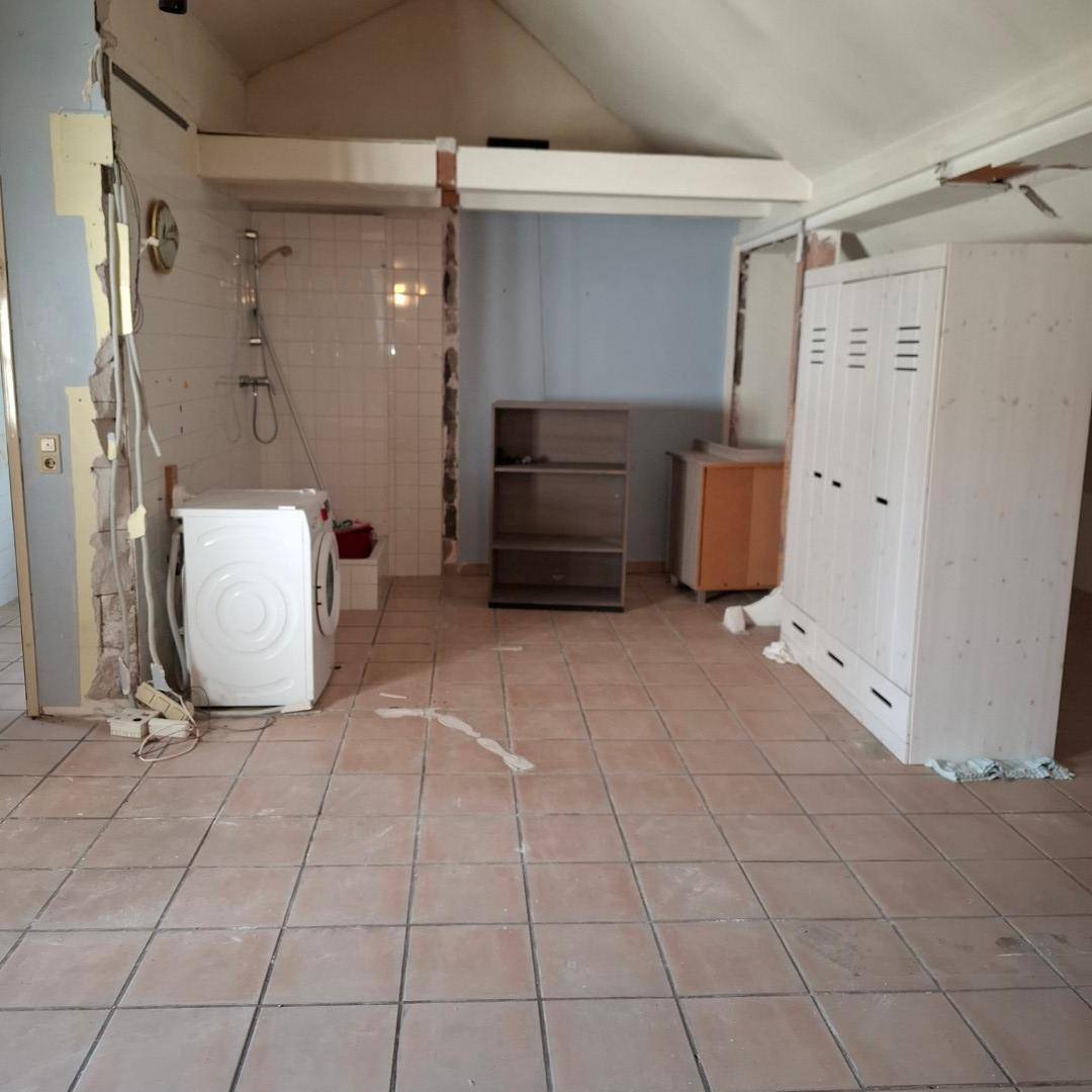 Raum in Renovierung mit Waschmaschine, Duschbereich und Möbeln, der Boden ist gefliest.