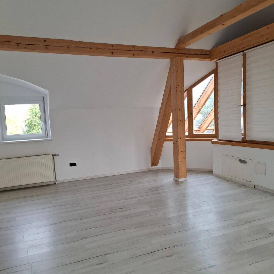 Heller Wohnraum mit Holzdielenboden, sichtbaren Holzbalken und großen Fenstern mit Jalousien unter Dachschrägen.