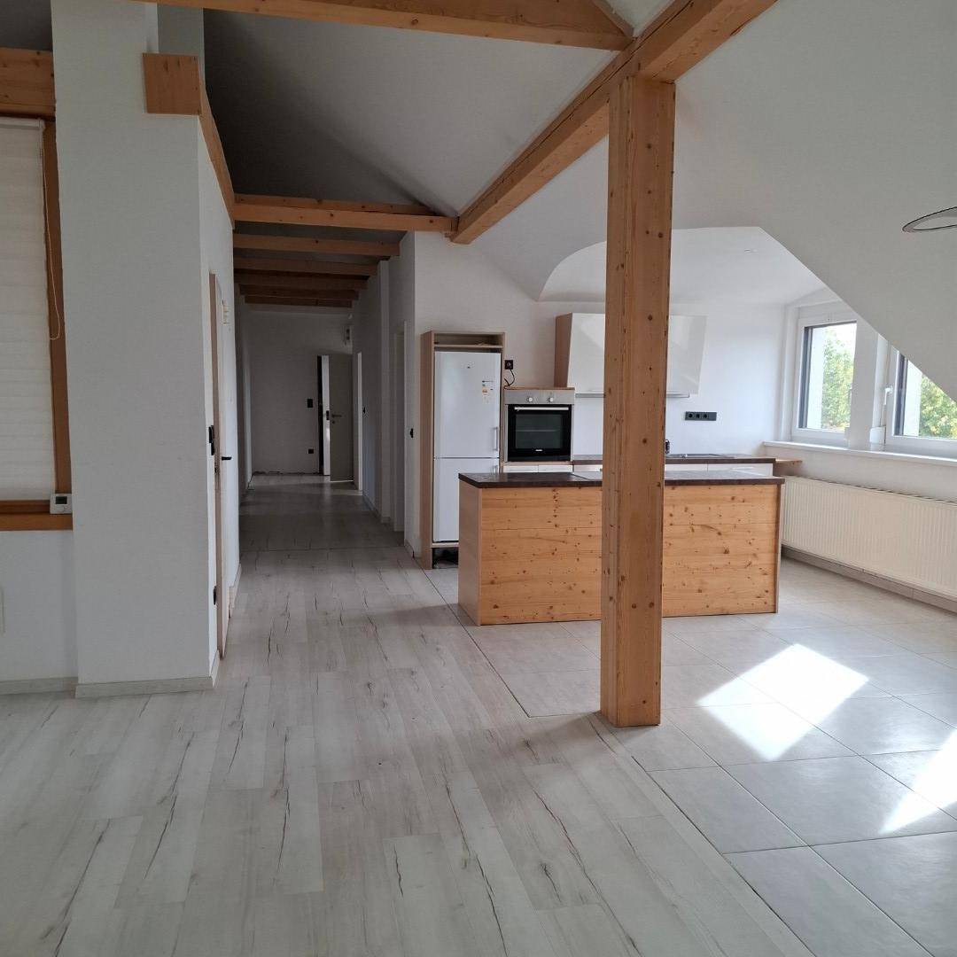 Offener Wohnbereich mit Küchenzeile, hellem Laminatboden und sichtbaren Holzbalken, mit Blick in den Flur.
