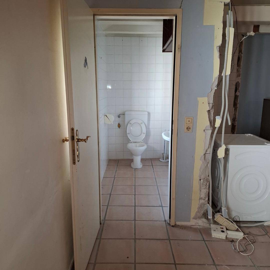 Blick durch eine Tür in ein kleines WC mit wandhängendem WC und Waschbecken, umgeben von Renovierungsarbeiten.