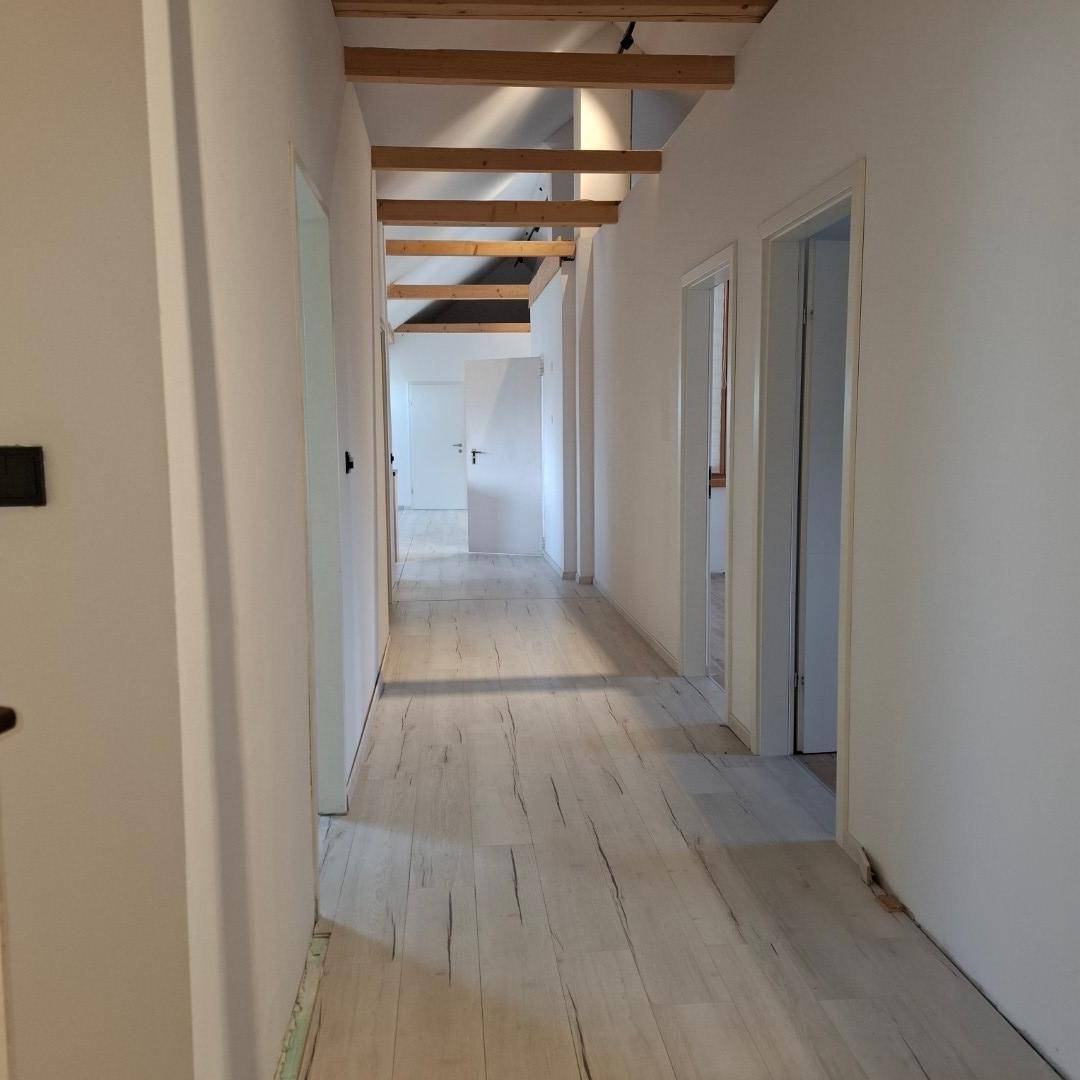 Heller Flur mit hellem Laminatboden, weißen Wänden und sichtbaren Holzdeckenbalken, der zu weiteren Räumen führt.