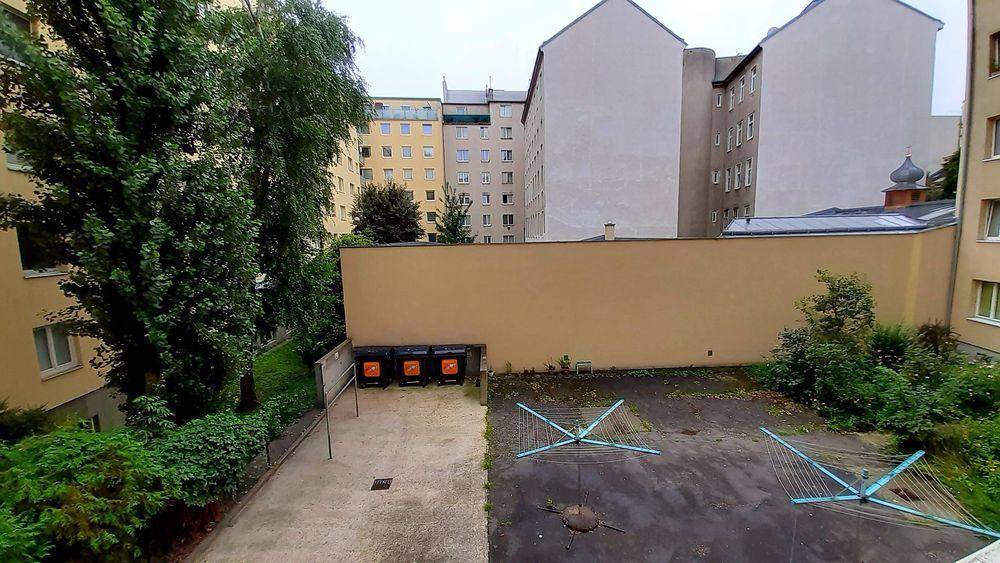 Blick auf einen Innenhof mit Bäumen, Mülltonnen und Wäscheständern, umgeben von Gebäuden.