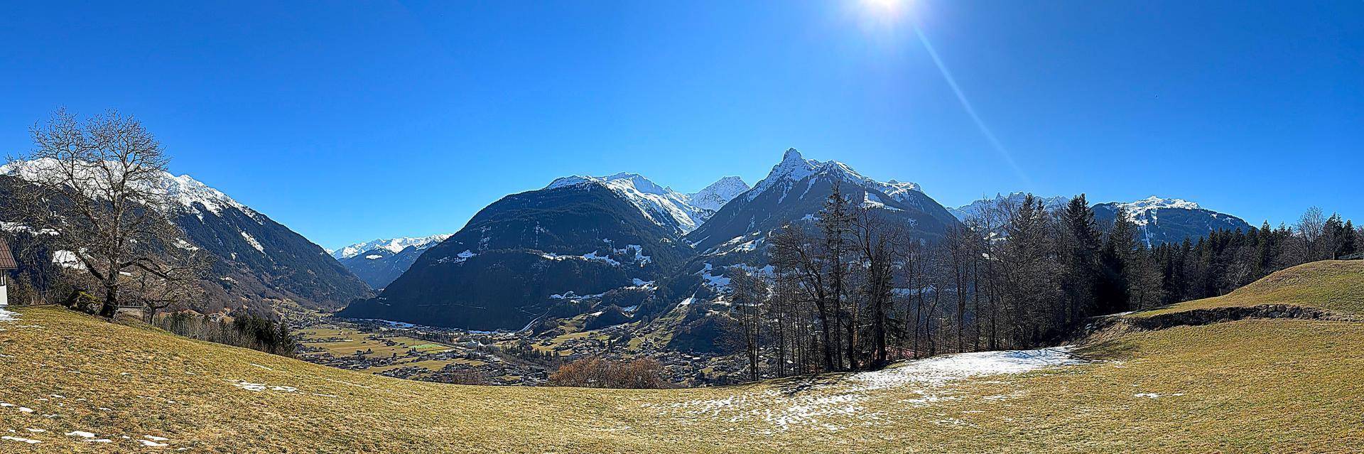 Weitläufiges Panorama der schneebedeckten Alpen und eines grünen Tals unter strahlend blauem Himmel.