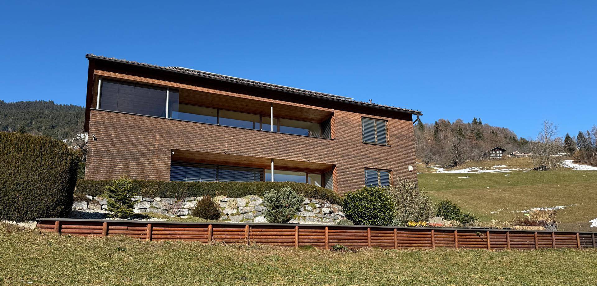 Moderne Architektur des Hauses mit großen Fenstern und einer gepflegten Gartenanlage am Hang.