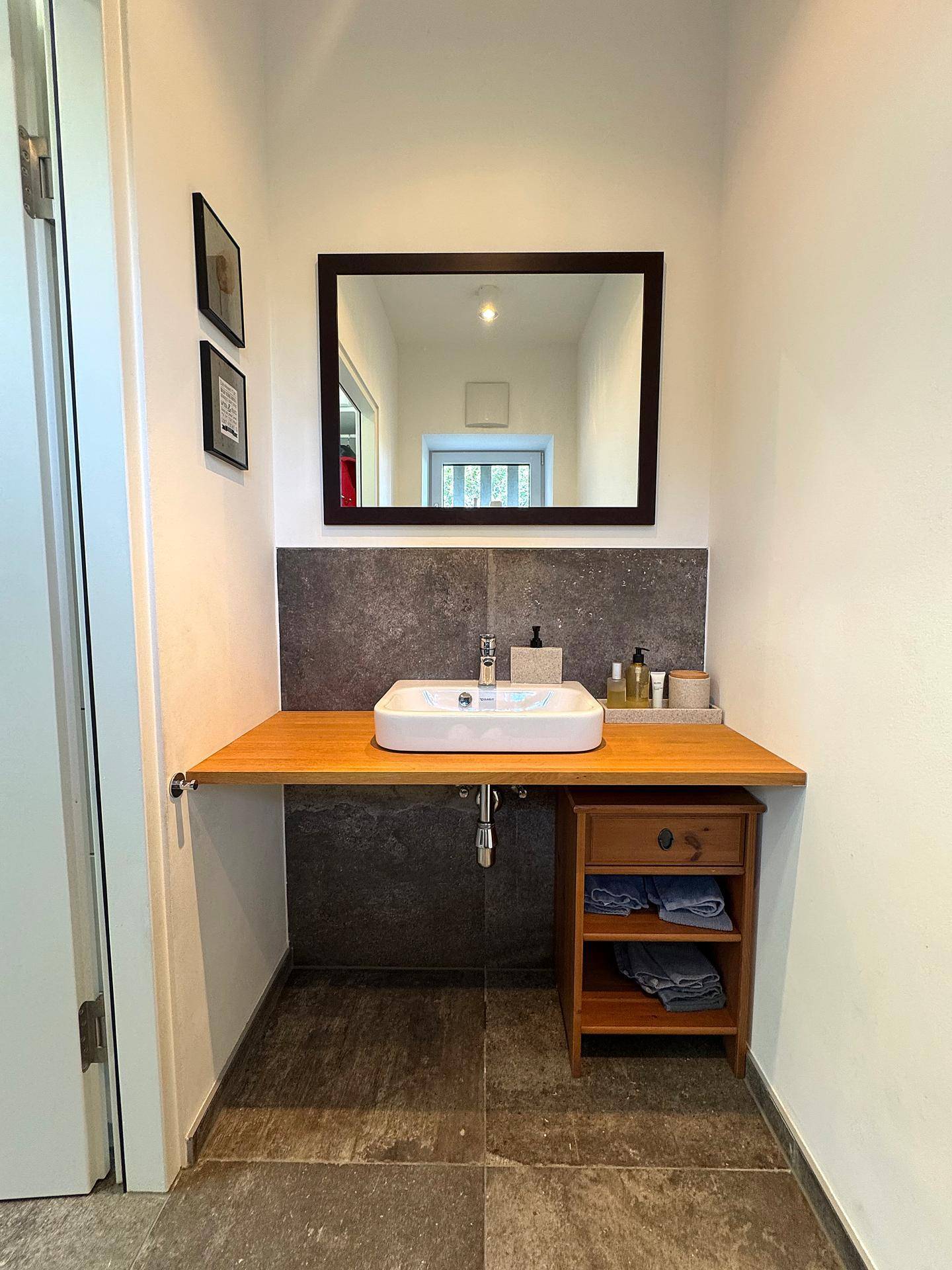 Kleines Badezimmer oder Gäste-WC mit Einzelwaschbecken, Holzablage und dunklen Fliesen.