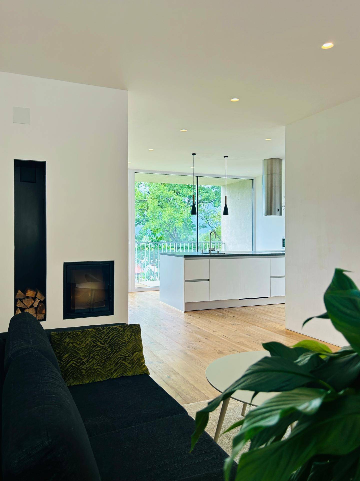 Gemütliches Wohnzimmer mit modernem Kamin, Holzboden und offenem Blick zur Küche und den Bergen.