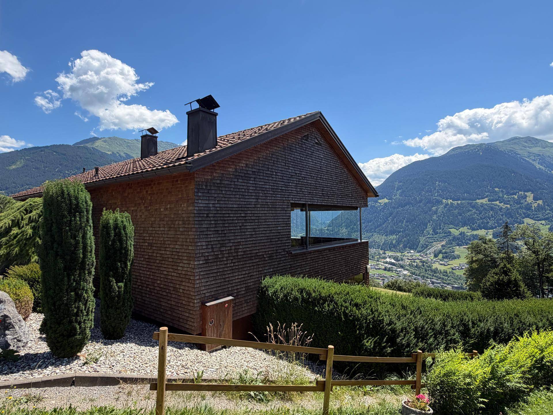 Seitenansicht des Hauses mit Holzfassade und großen Fenstern, eingebettet in die Berglandschaft.