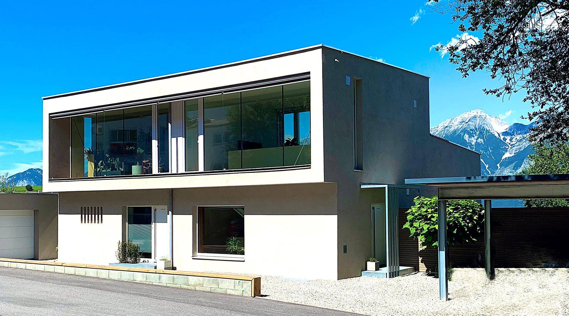 Modernes Wohnhaus mit großer Glasfassade und Bergblick unter strahlend blauem Himmel.