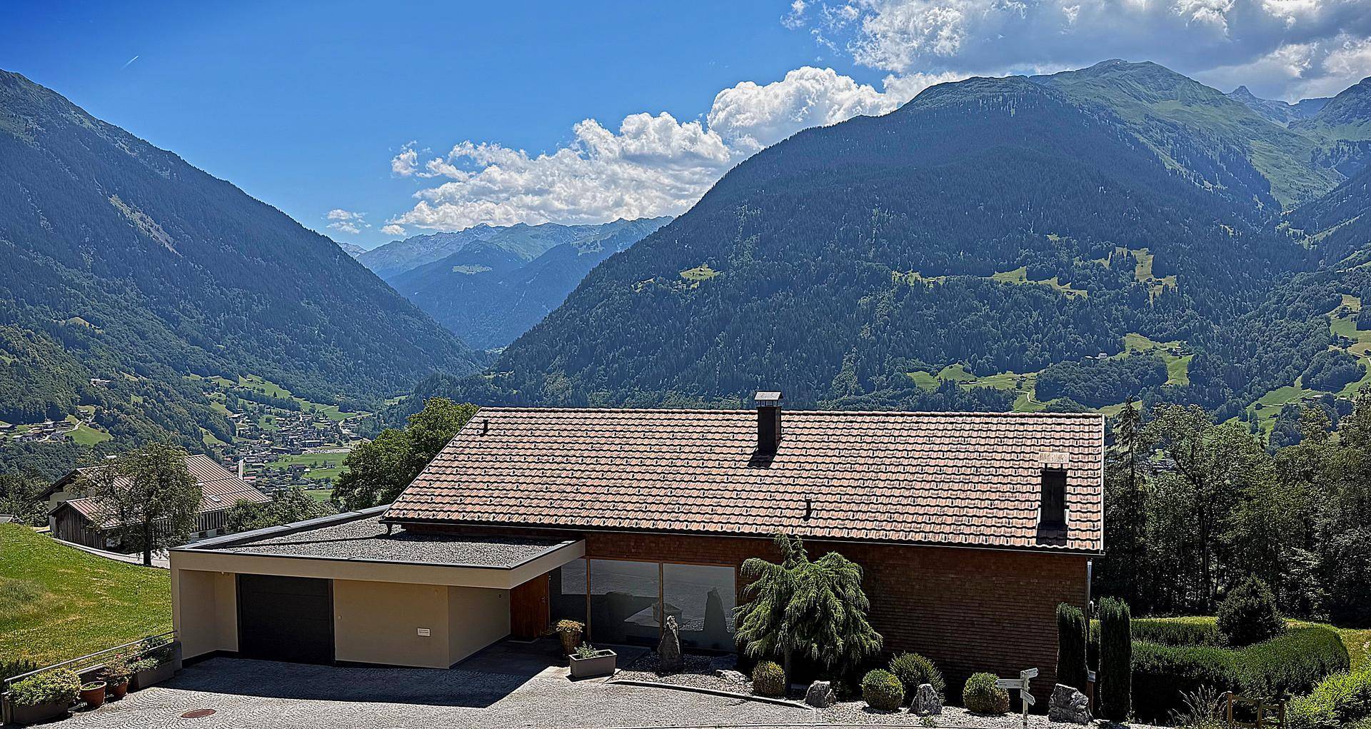 Haus in alpiner Landschaft mit Garage und gepflegtem Garten, umgeben von grünen Bergen.