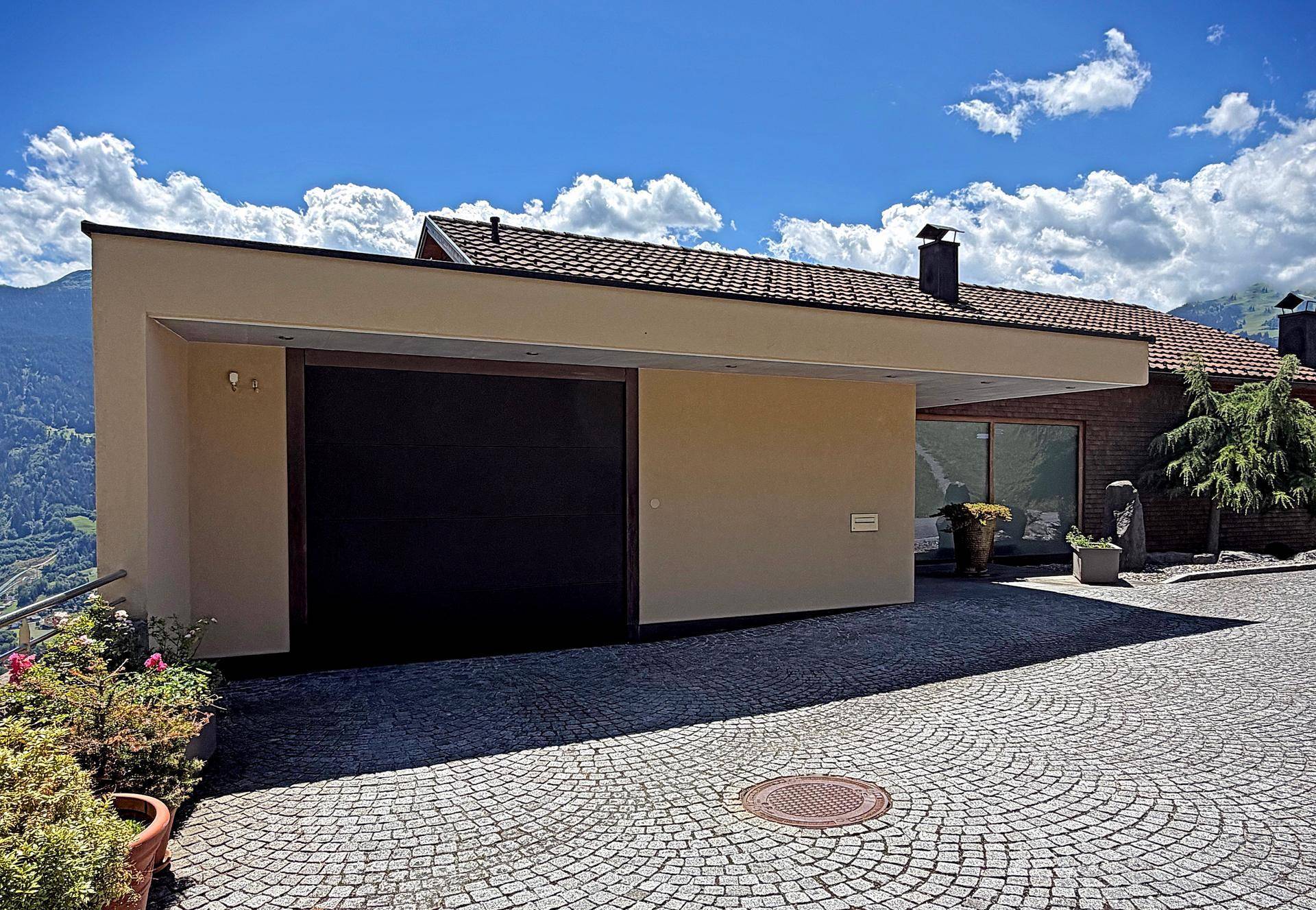 Moderne Garage mit flachem Dach und gepflasterter Auffahrt, umgeben von alpiner Natur.
