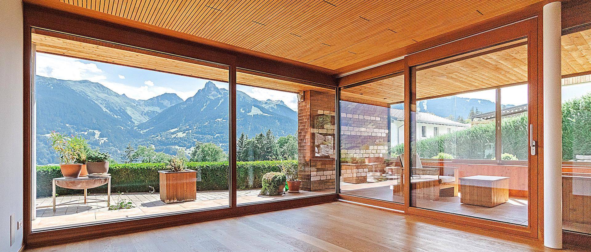 Lichtdurchfluteter Raum mit Holzboden, großen Fenstern und Zugang zu einer Terrasse mit Bergblick.