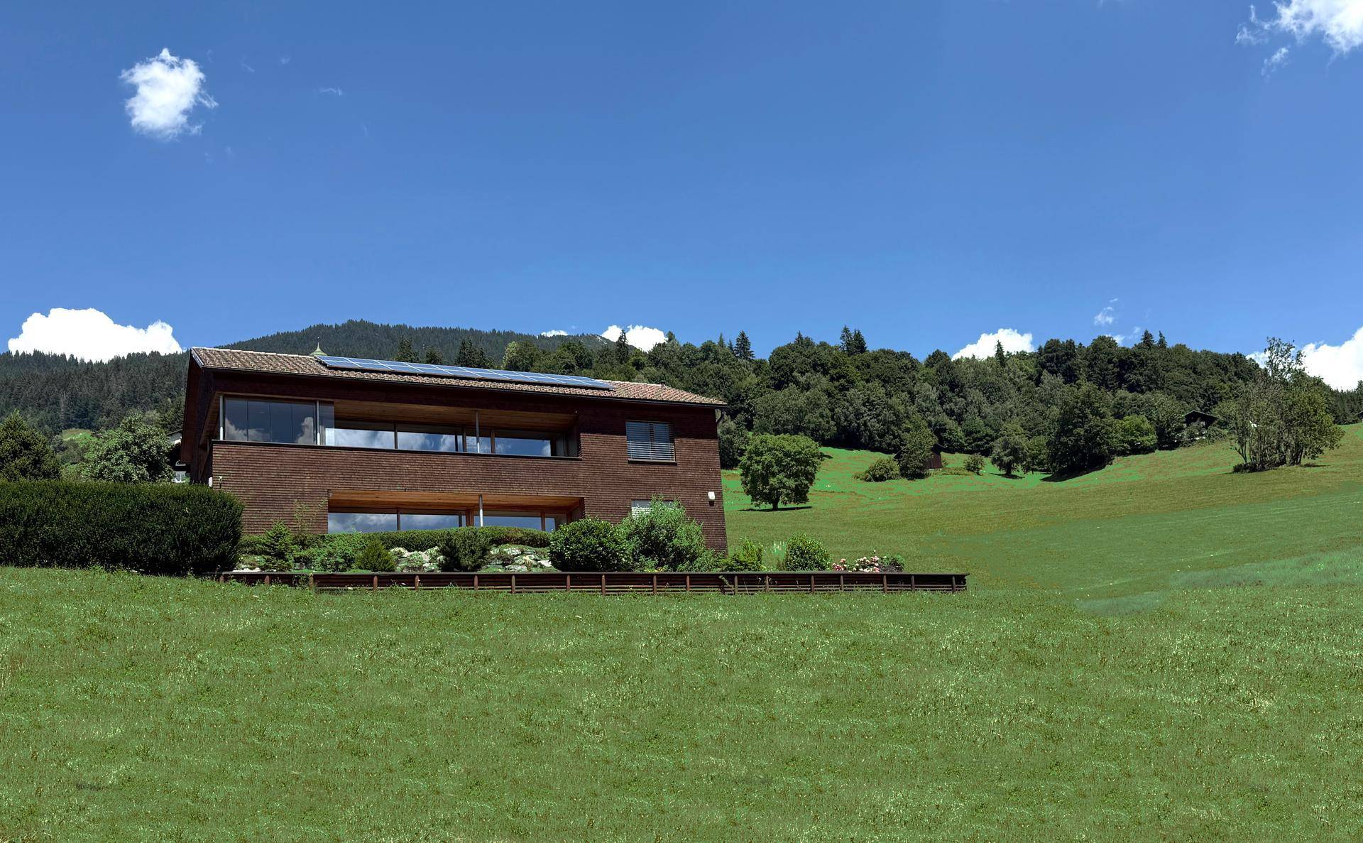 Modernes Haus mit Photovoltaikanlage auf dem Dach, umgeben von grünen Wiesen und Bergen im Sommer.