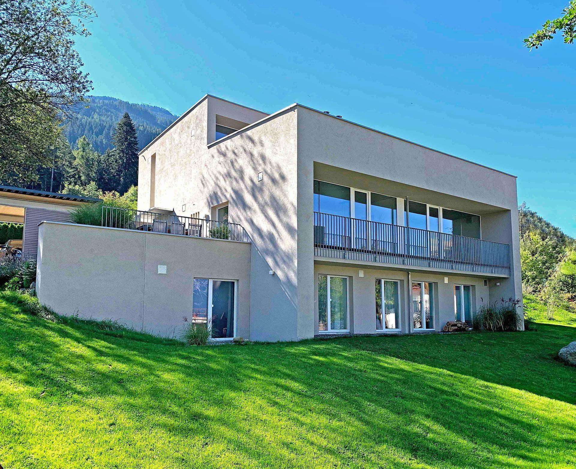Modernes Haus mit weitläufigem grünen Garten und großen Fenstern in alpiner Umgebung.