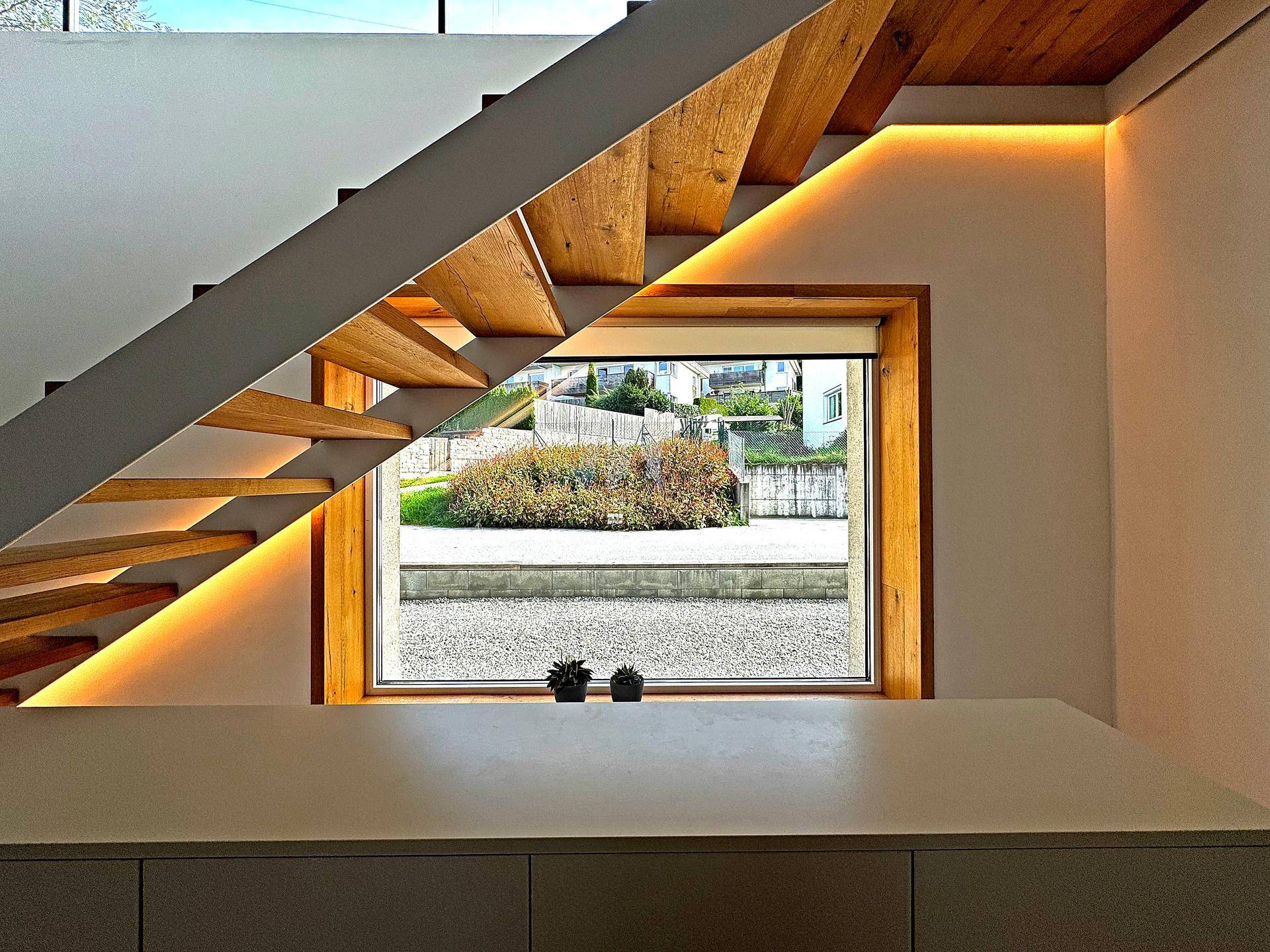 Detailansicht einer modernen Holztreppe mit indirekter Beleuchtung und Fensterblick nach draußen.
