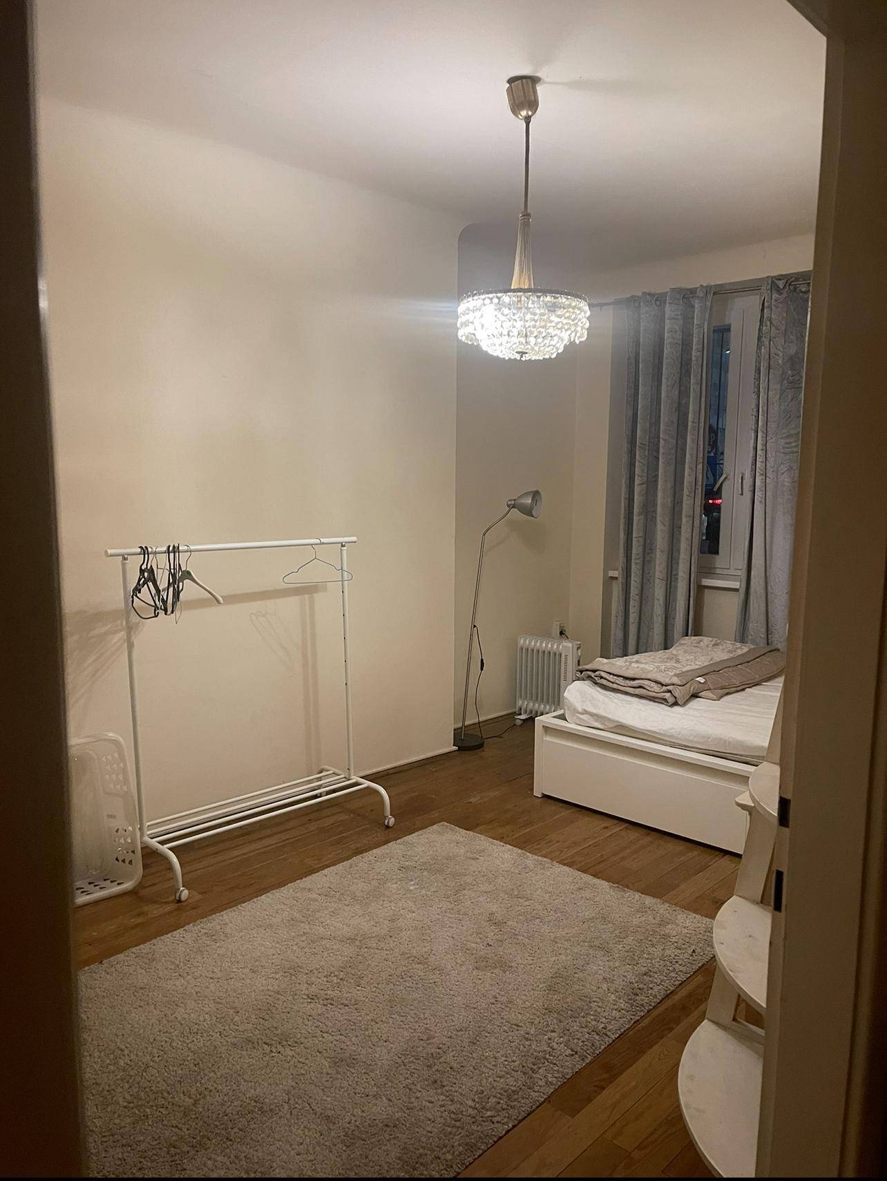 Schlafzimmer mit Holzboden, Bett, Kleiderständer und einem modernen Kronleuchter.