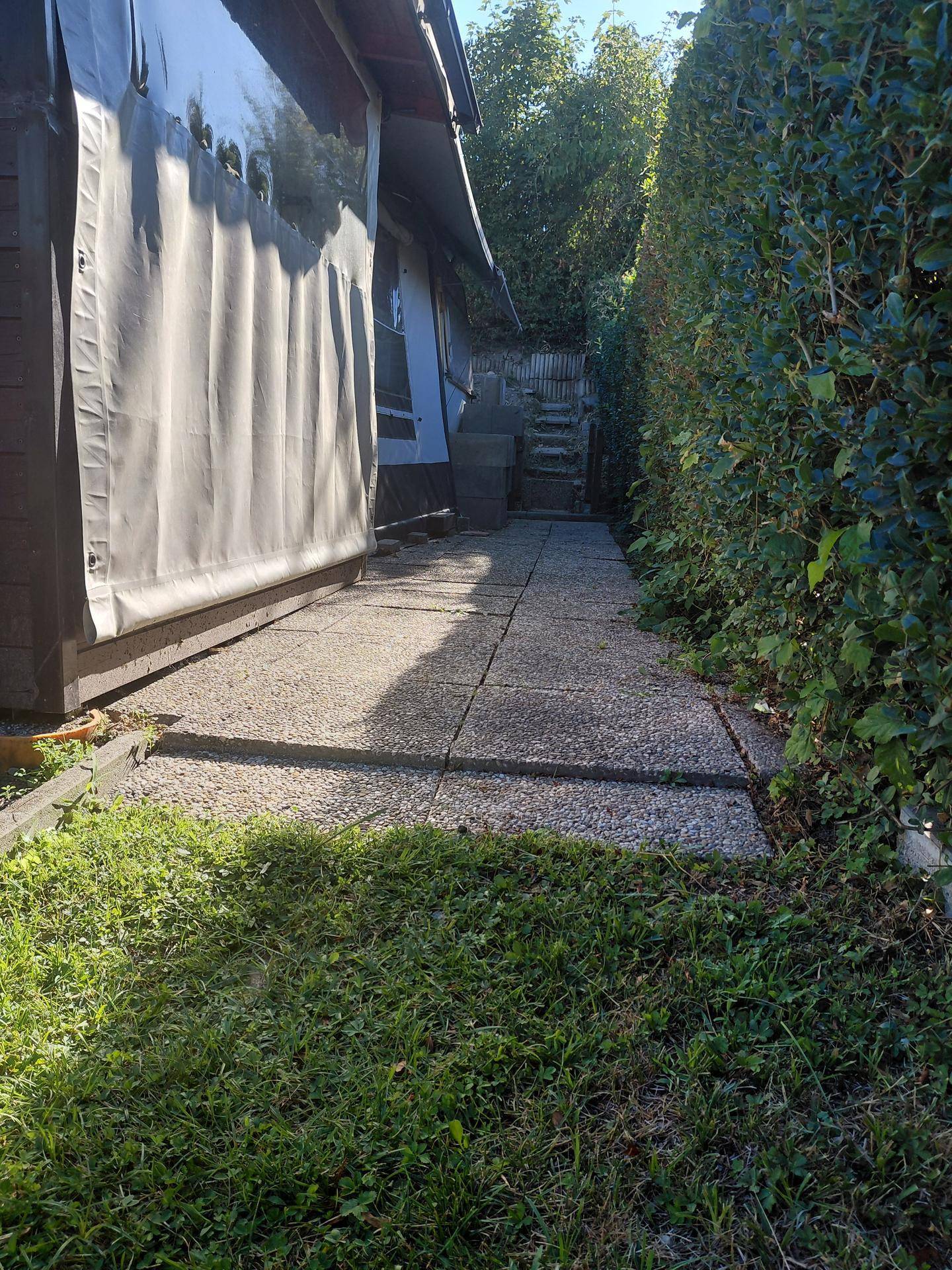 Schmaler gepflasterter Weg entlang eines Gebäudes und einer hohen, dichten Hecke im Freien.