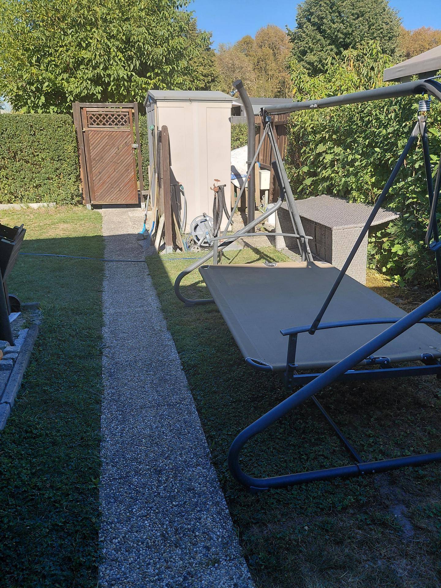 Gartenbereich mit einer Hollywoodschaukel, einem Schuppen und einem Holztor im Hintergrund.