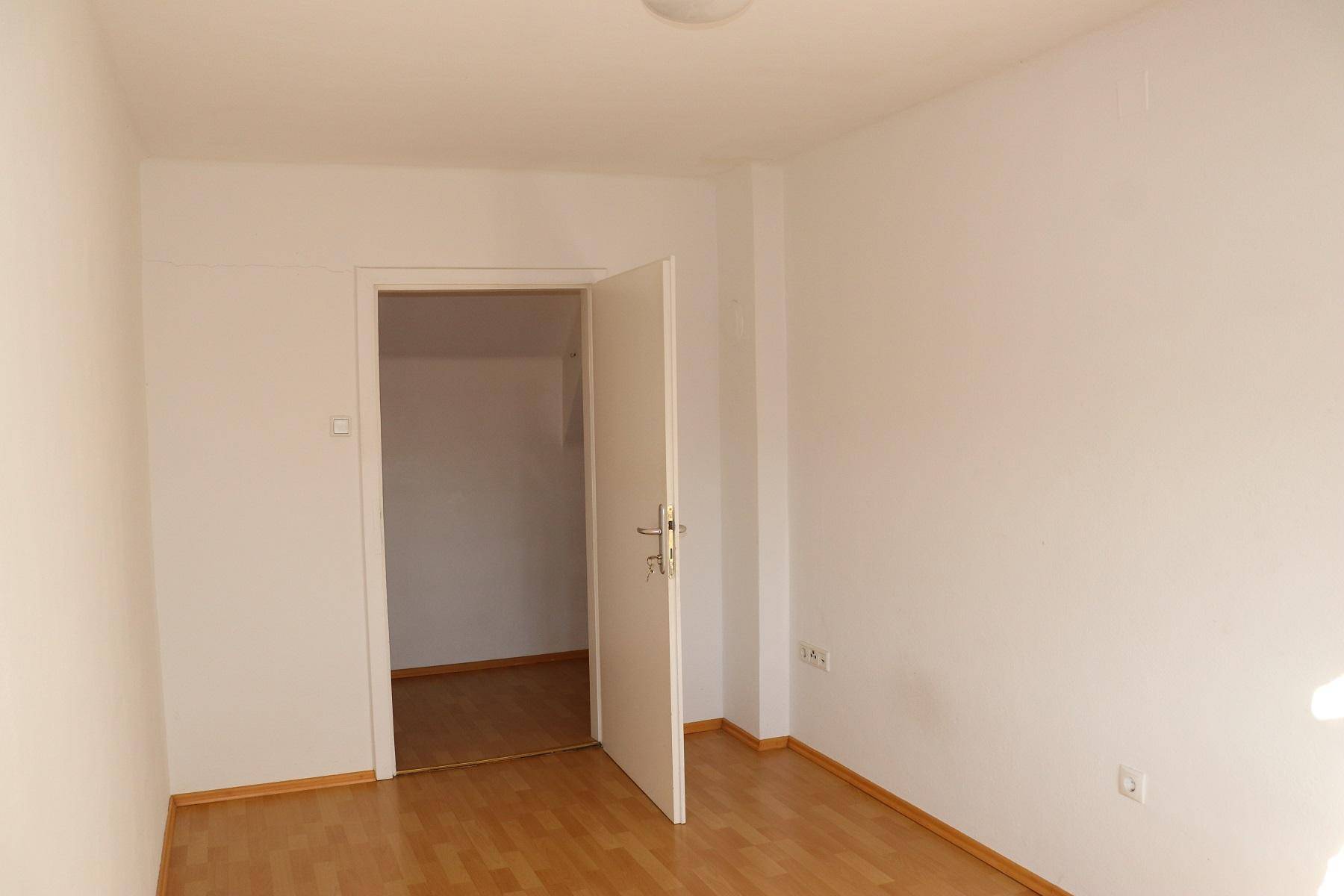 Leerer Raum mit Laminatboden und offener Tür, die den Blick in ein weiteres Zimmer freigibt.