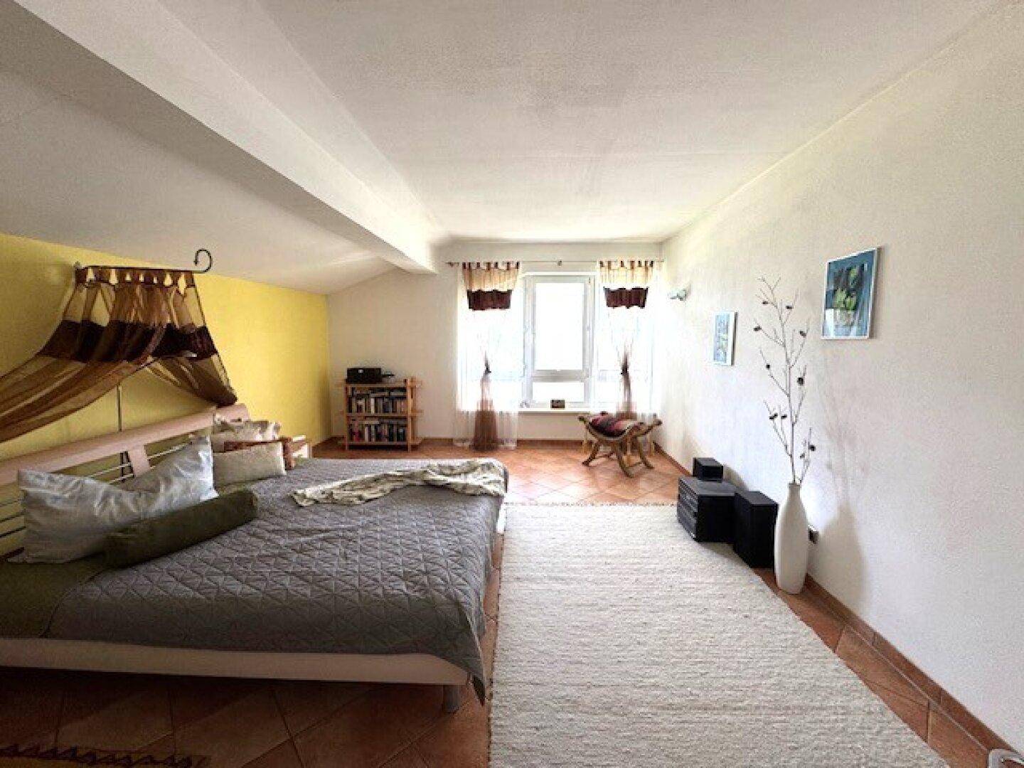 Geräumiges Schlafzimmer mit Terracottaboden, Teppich, gelber Akzentwand und Fenster.