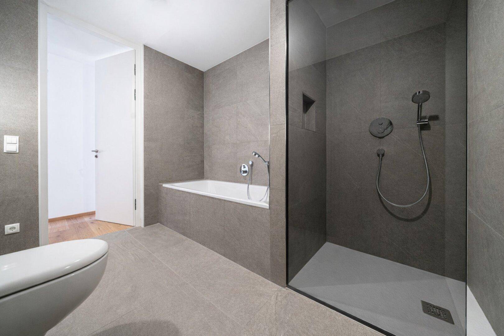 Modernes Badezimmer mit grauen Fliesen, Badewanne, ebenerdiger Dusche und wandhängendem WC.