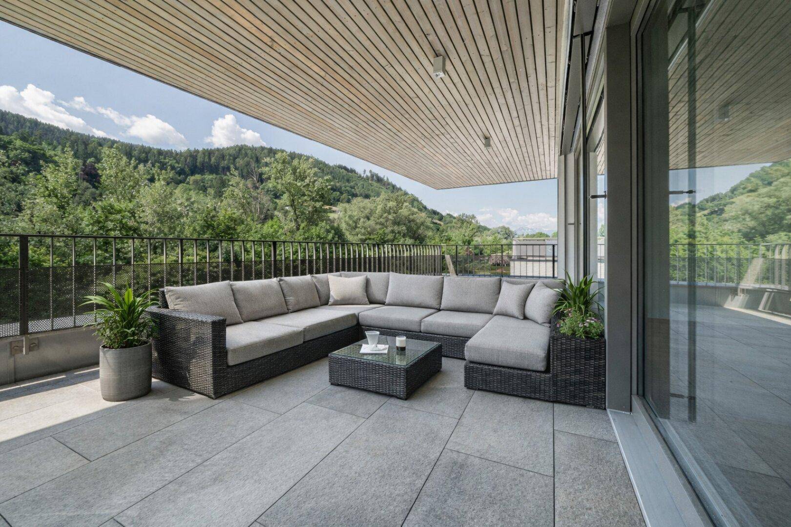 Große überdachte Terrasse mit bequemen Loungemöbeln, Fliesenboden und Panoramablick auf die bewaldeten Berge.