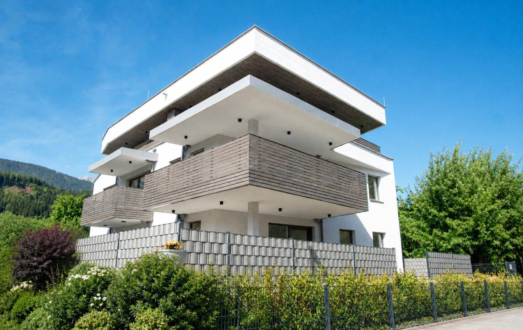 Modernes Mehrfamilienhaus mit mehreren Balkonen und gepflegter Gartenanlage unter blauem Himmel.