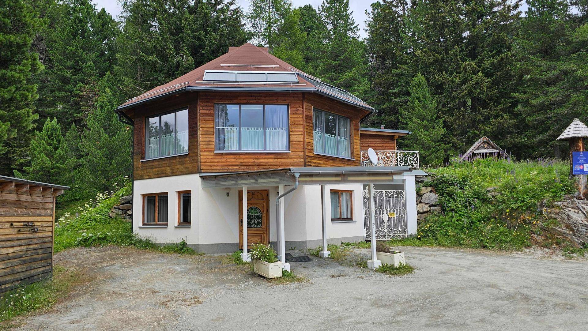 Charmantes achteckiges Haus mit Holzverkleidung und weißem Sockel, umgeben von üppiger Natur und Bäumen.