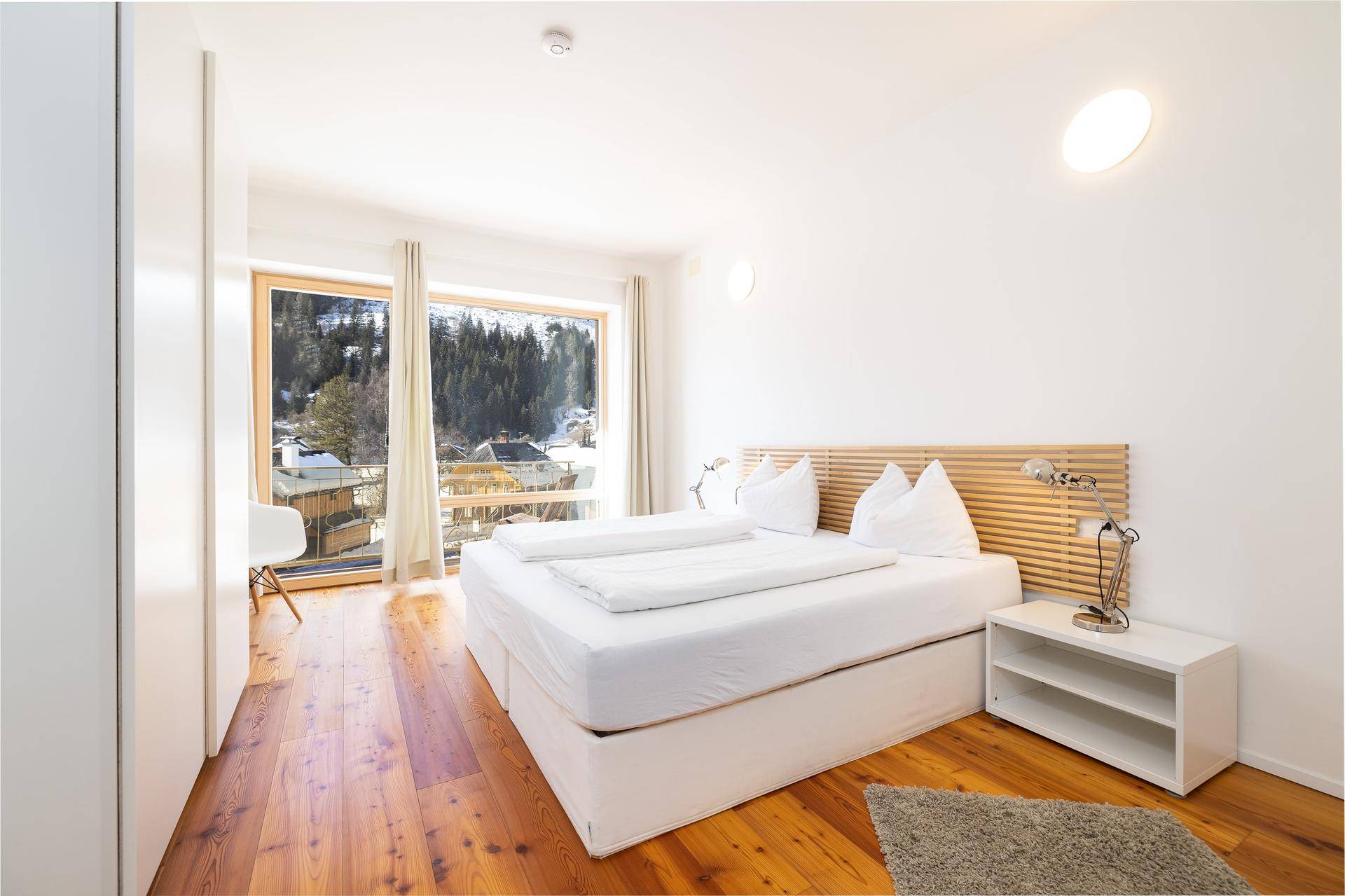 Helles Schlafzimmer mit weißem Bett, Holzboden und großen Fenstern mit Blick auf die verschneite Natur.