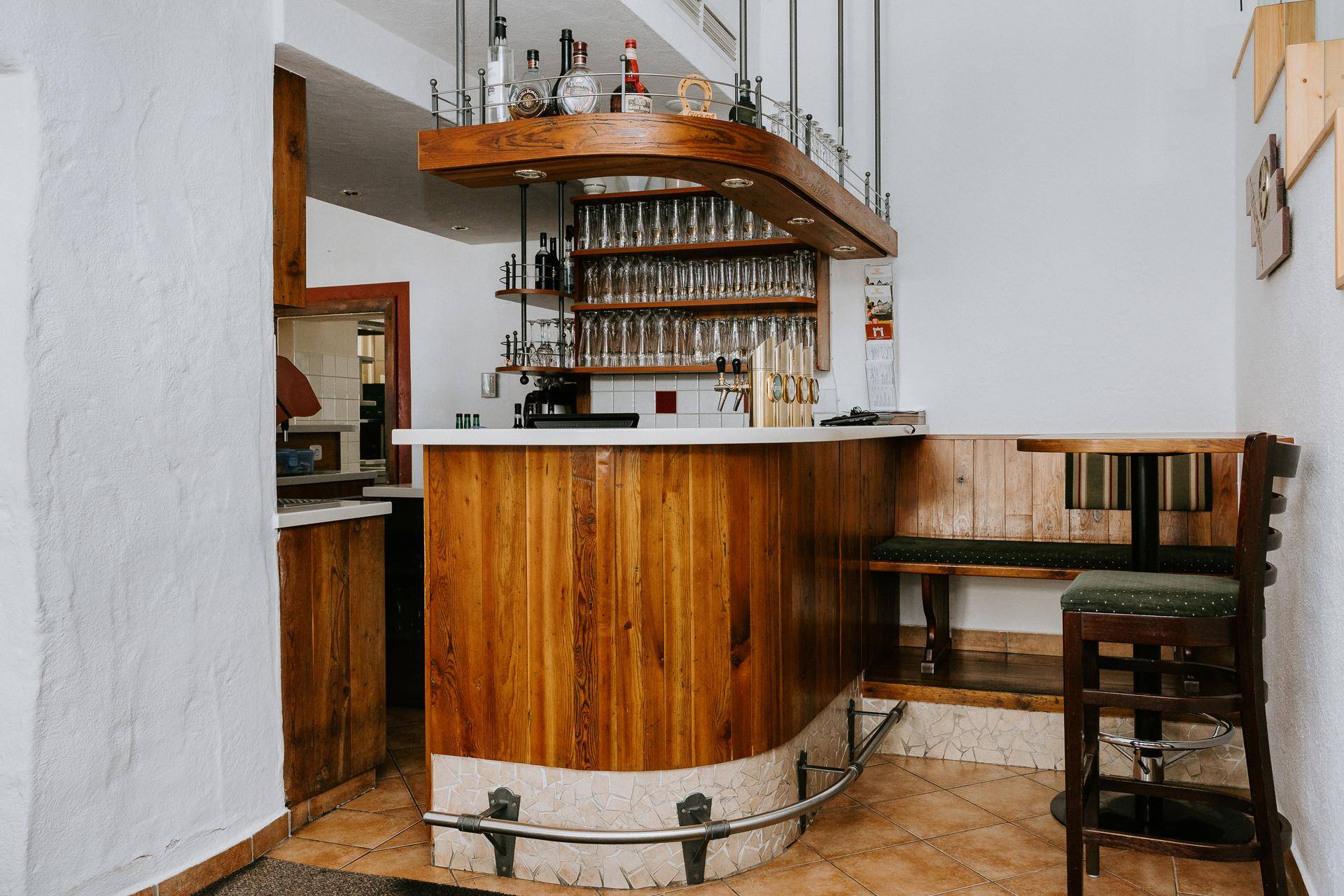 Rustikale Bar mit Holztheke, Fliesenboden und Regalen für Gläser und Flaschen.