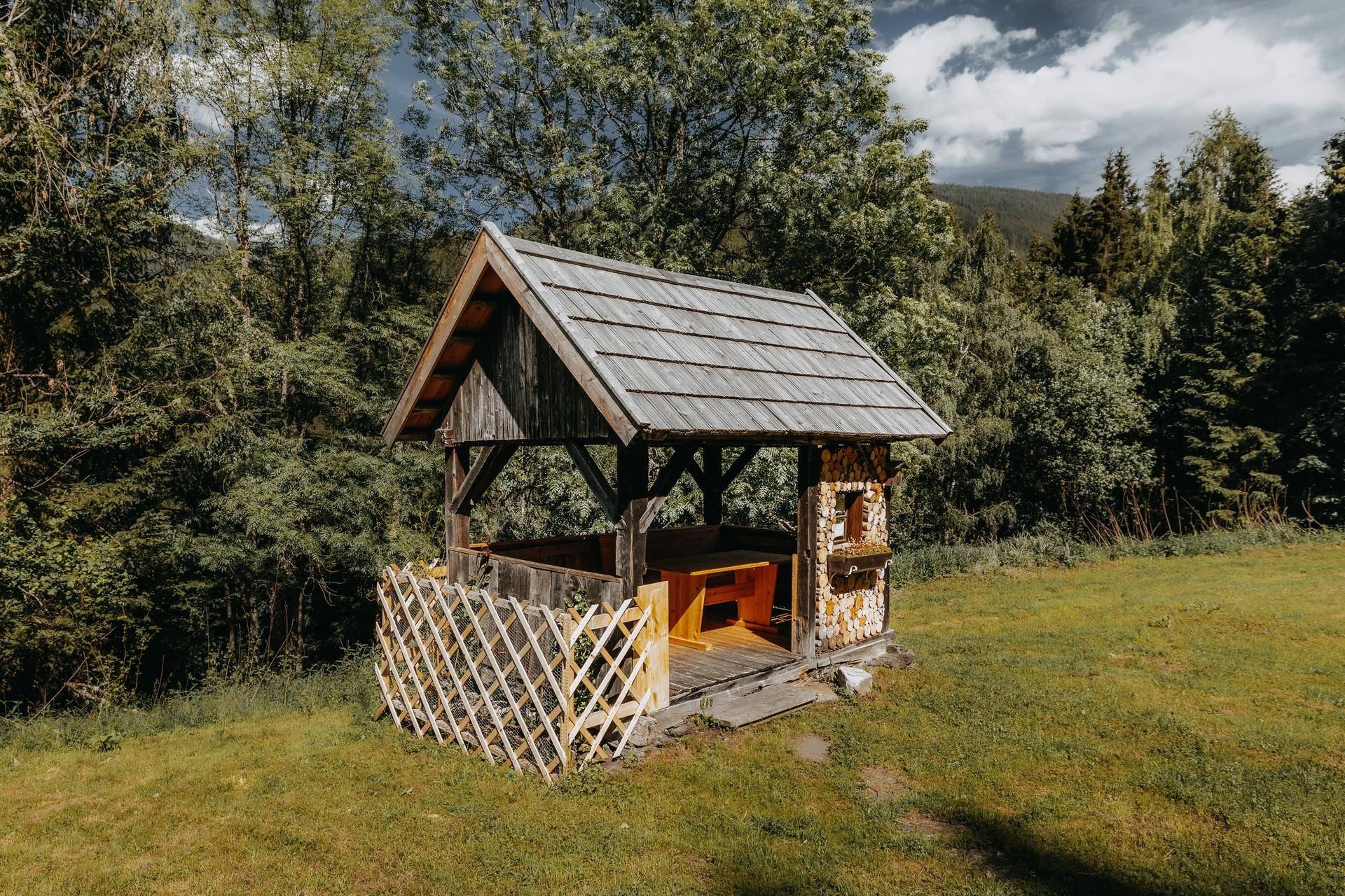Gemütlicher Holzpavillon mit Sitzgelegenheiten im Garten, umgeben von grüner Natur.