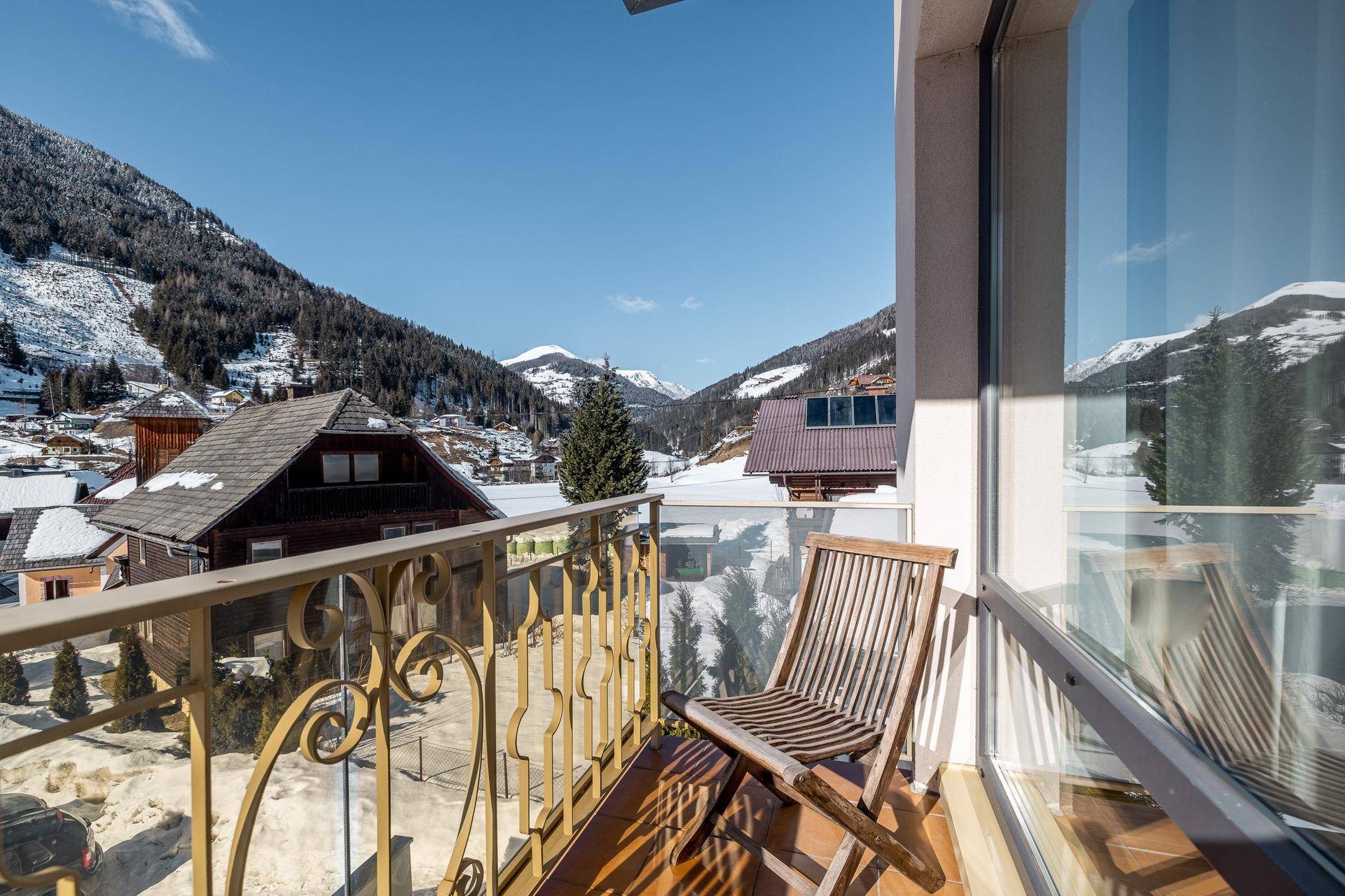 Kleiner Balkon mit Holzstuhl und Metallgeländer, bietet Blick auf ein verschneites Dorf und Berge.