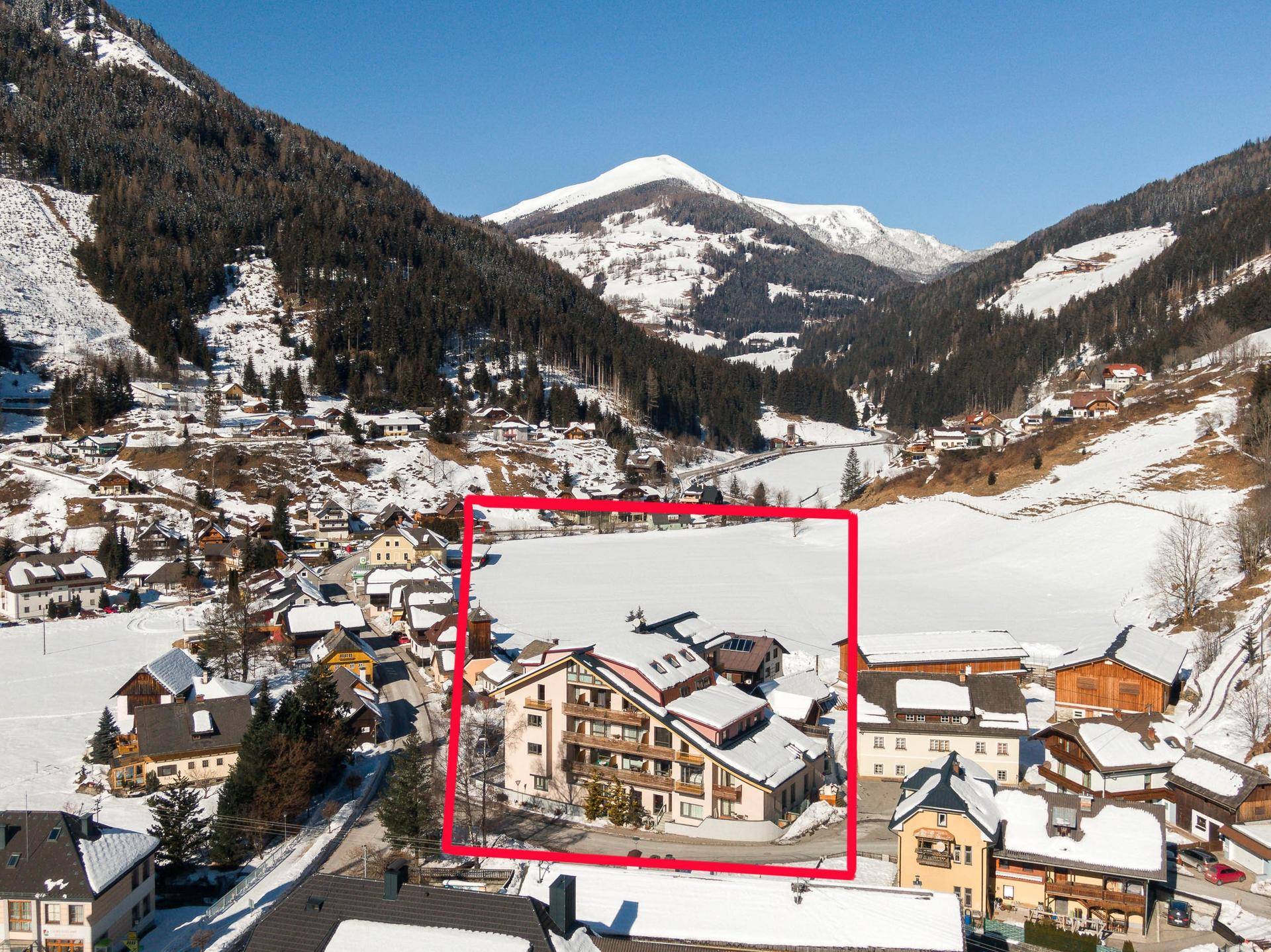 Luftaufnahme eines verschneiten Dorfes in einem Tal mit Bergen, das Hauptgebäude ist rot umrandet.
