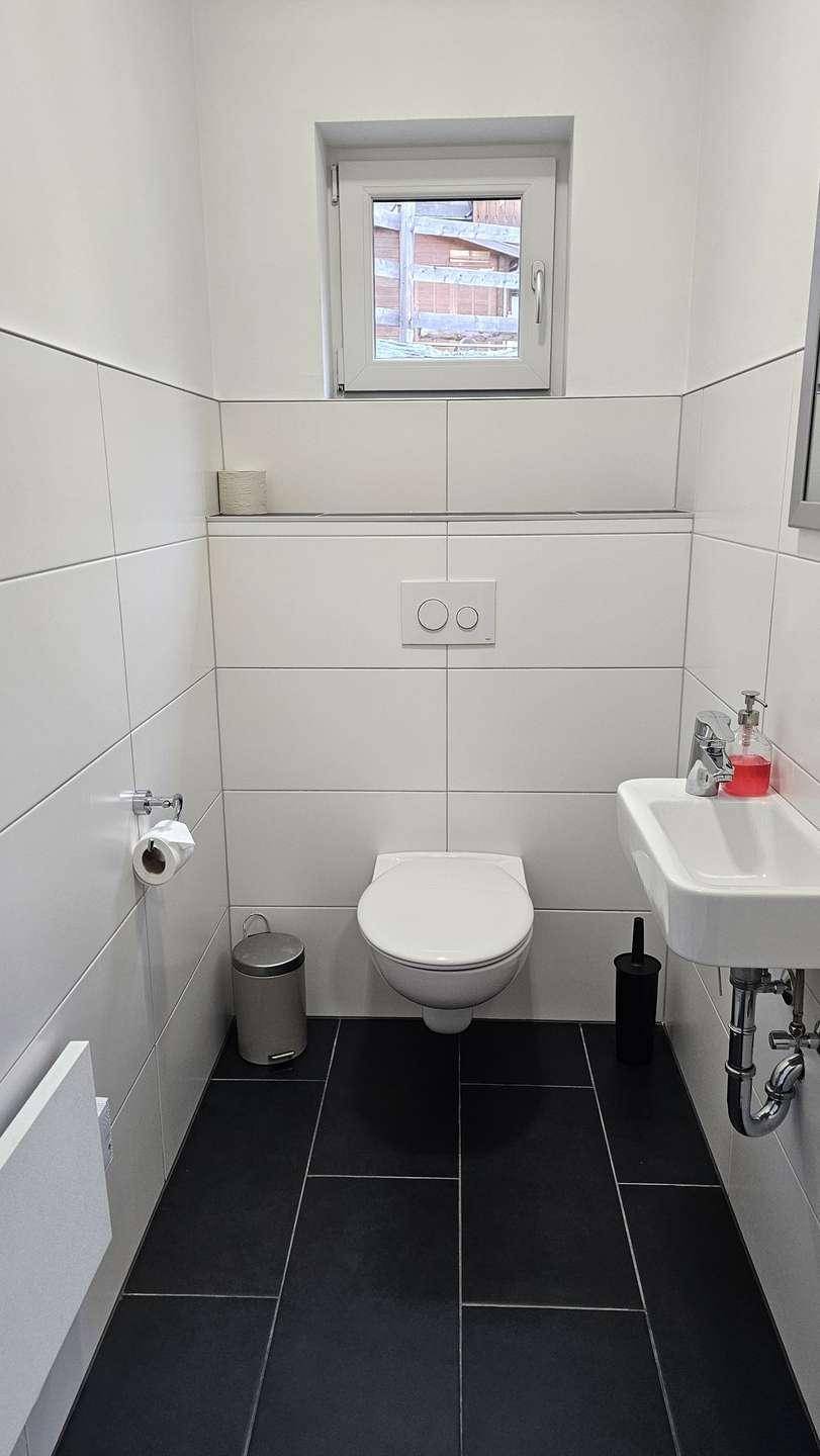 Helles Gäste-WC mit weißen Fliesen, dunklem Boden und einem Fenster für Tageslicht.