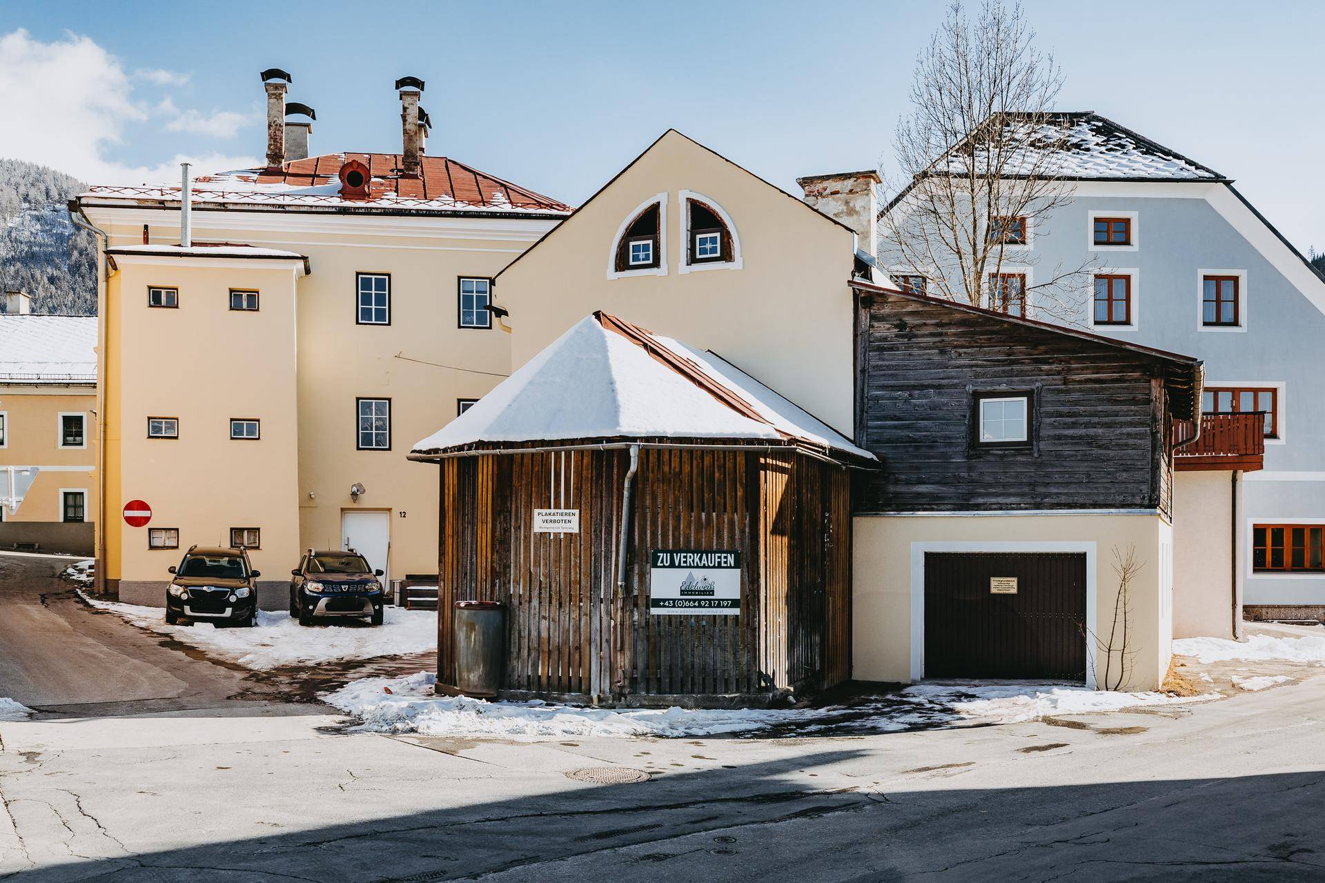 Mehrere Gebäude in einer verschneiten Gasse, darunter ein Holzschuppen mit Verkaufsanzeige.