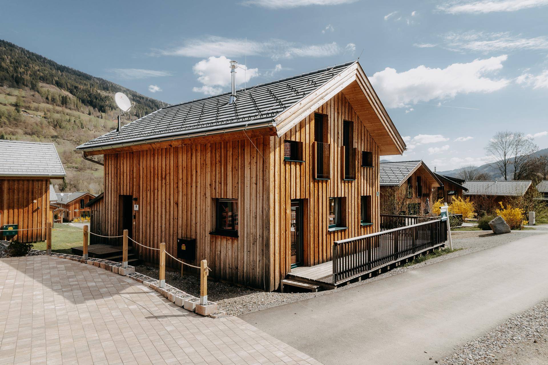 Modernes Holzhaus mit Satteldach und gepflastertem Zugangsweg in bergiger Landschaft.
