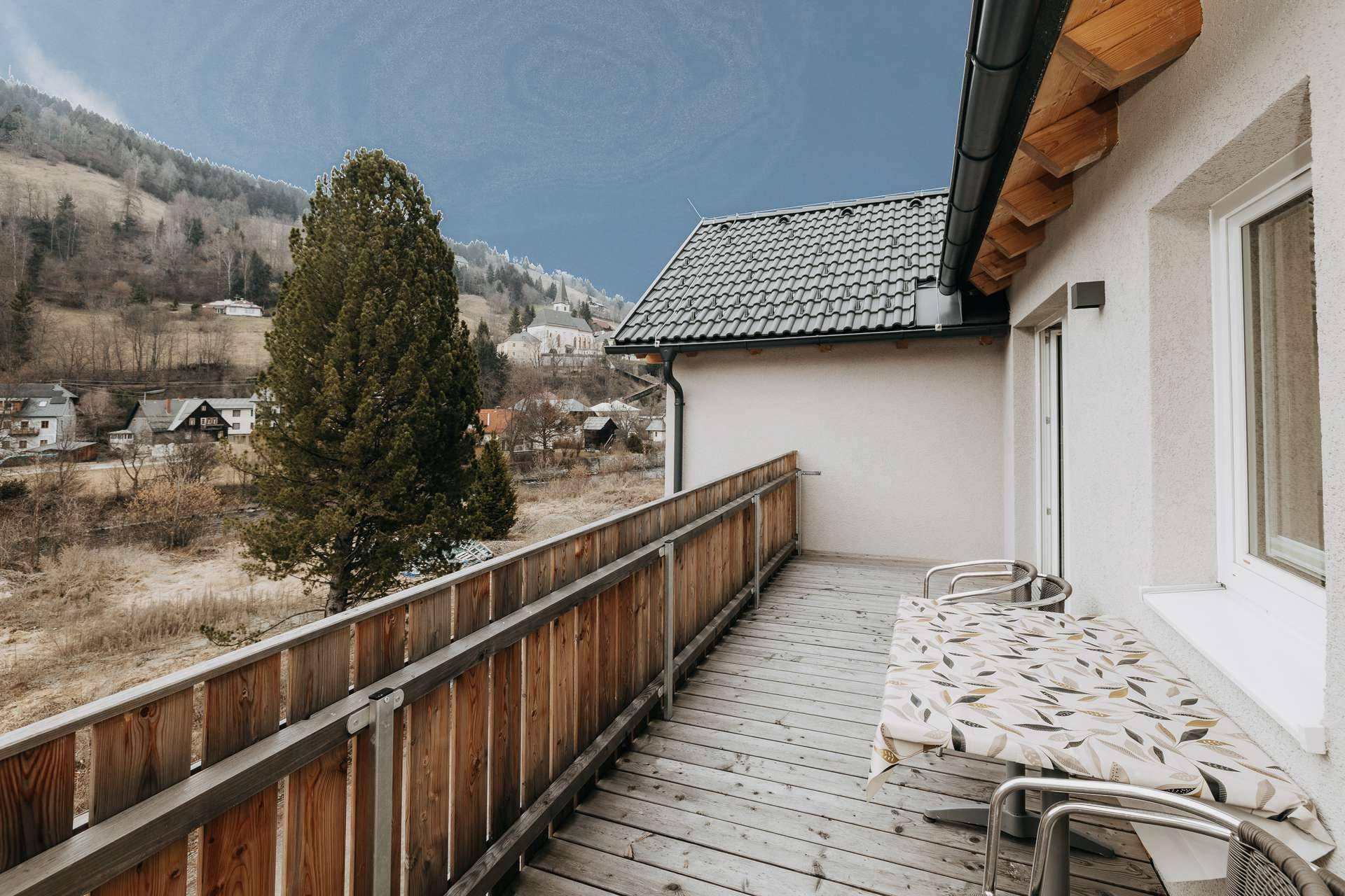 Geräumiger Holzbalkon mit Sitzgelegenheiten und weitem Blick auf die umliegende Berglandschaft und das Dorf.