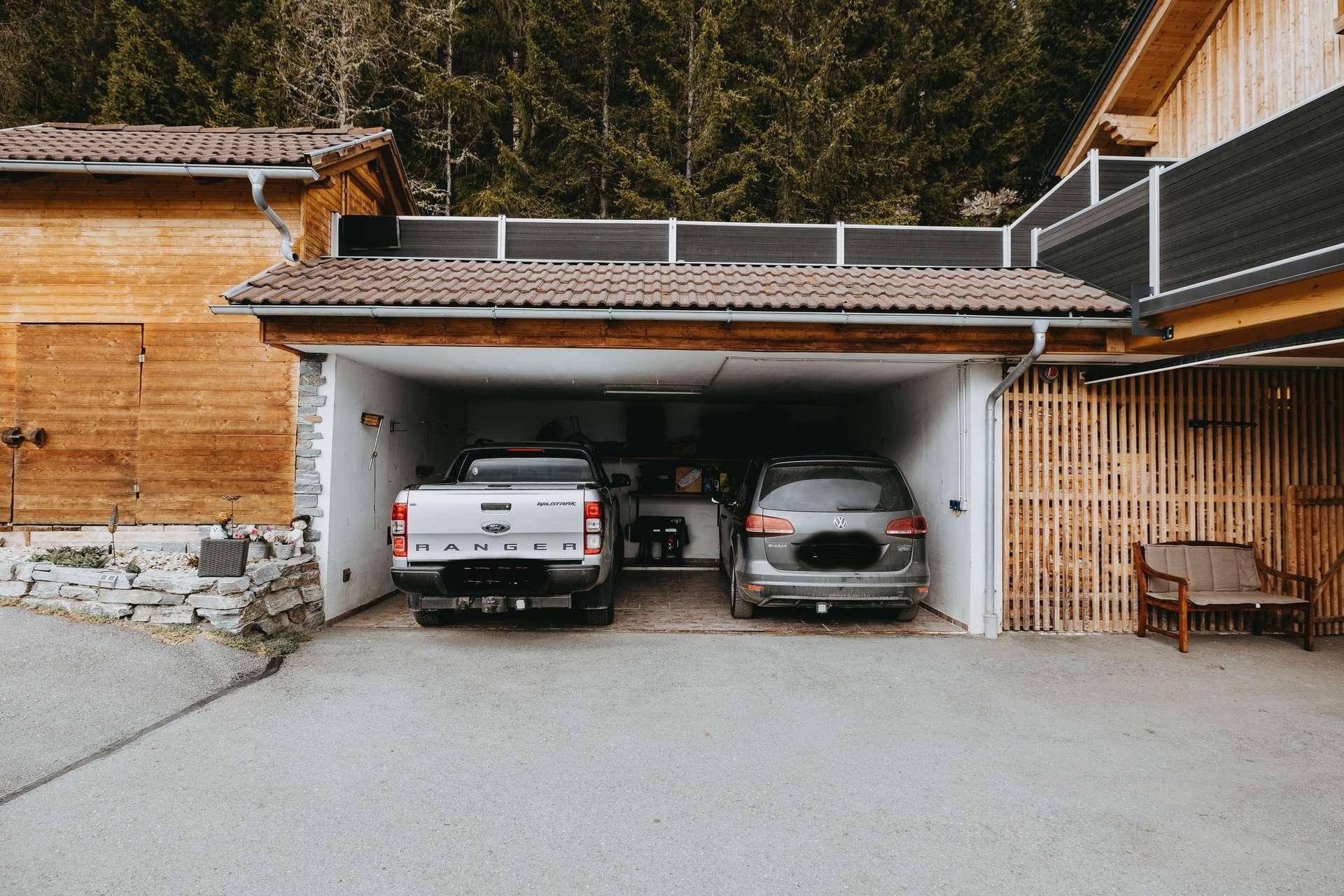 Doppelgarage mit zwei geparkten Fahrzeugen, umgeben von Holzelementen und einer Zufahrt.