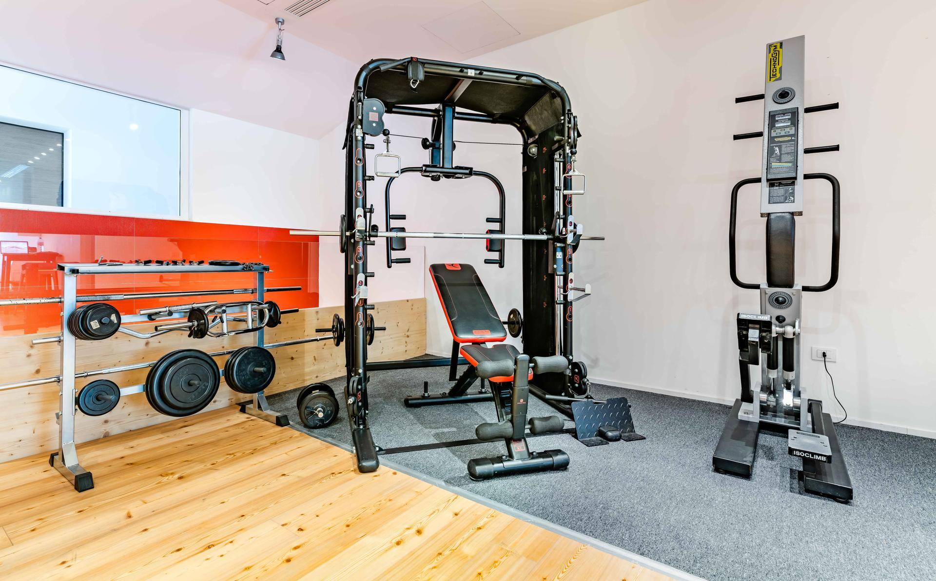 Fitnessraum mit Krafttrainingsgeräten, darunter eine Smith-Maschine und freie Gewichte, mit Holz- und Teppichboden.