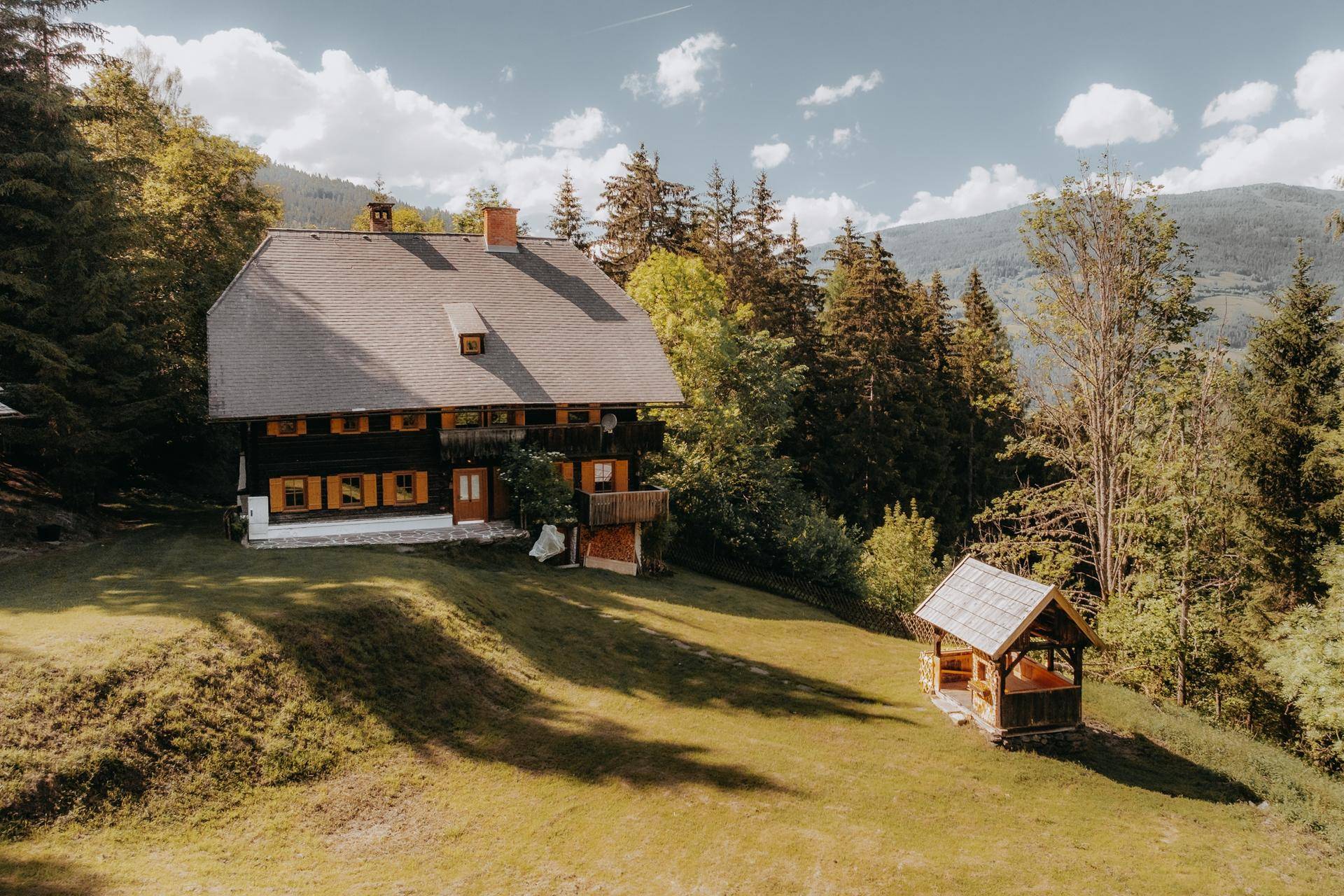 Traditionelles Holzhaus mit einem kleinen Holzpavillon auf einer weitläufigen grünen Wiese.