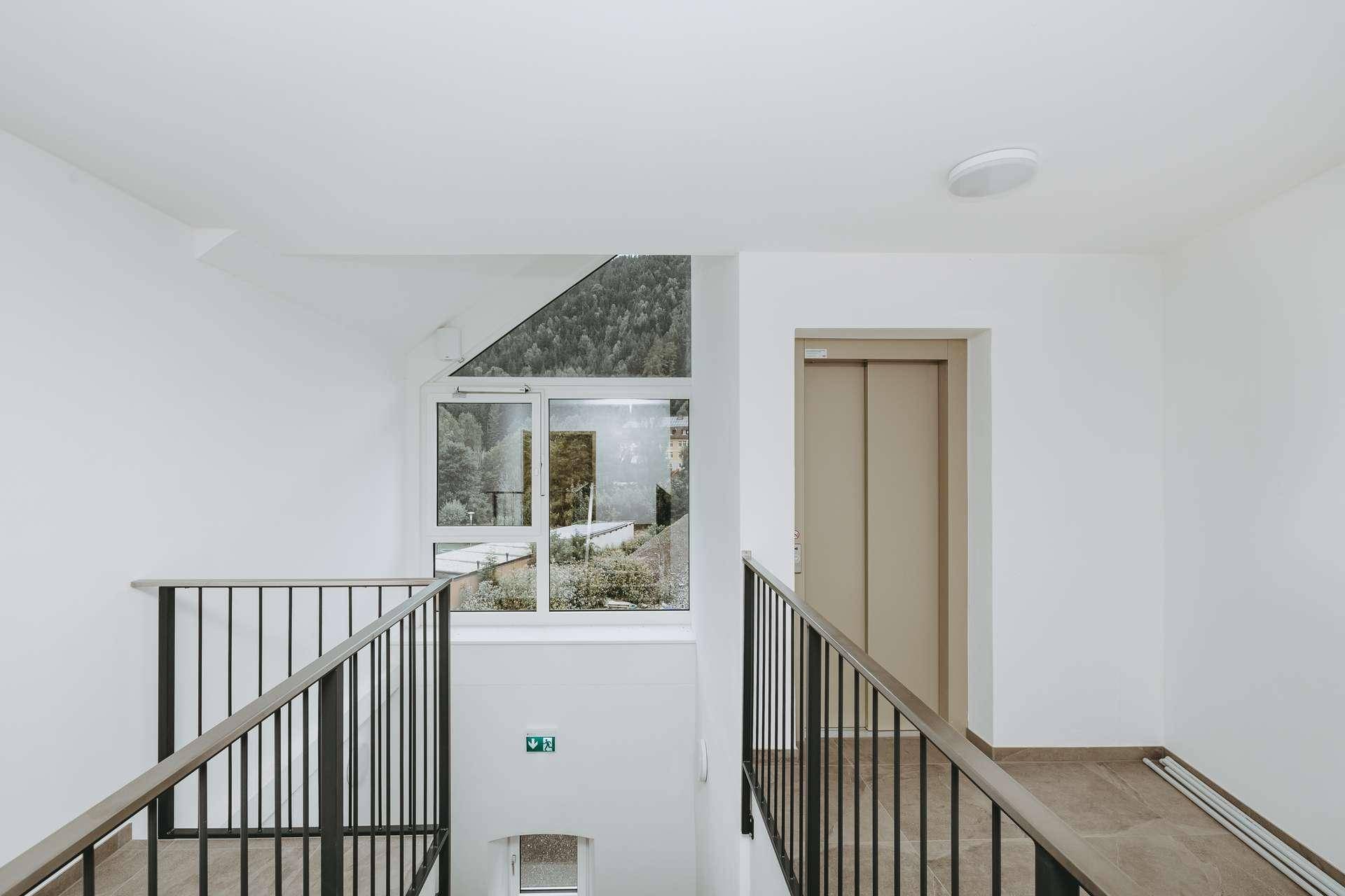 Helles Treppenhaus mit Geländer und Aufzug, bietet natürlichen Lichteinfall durch große Fenster.
