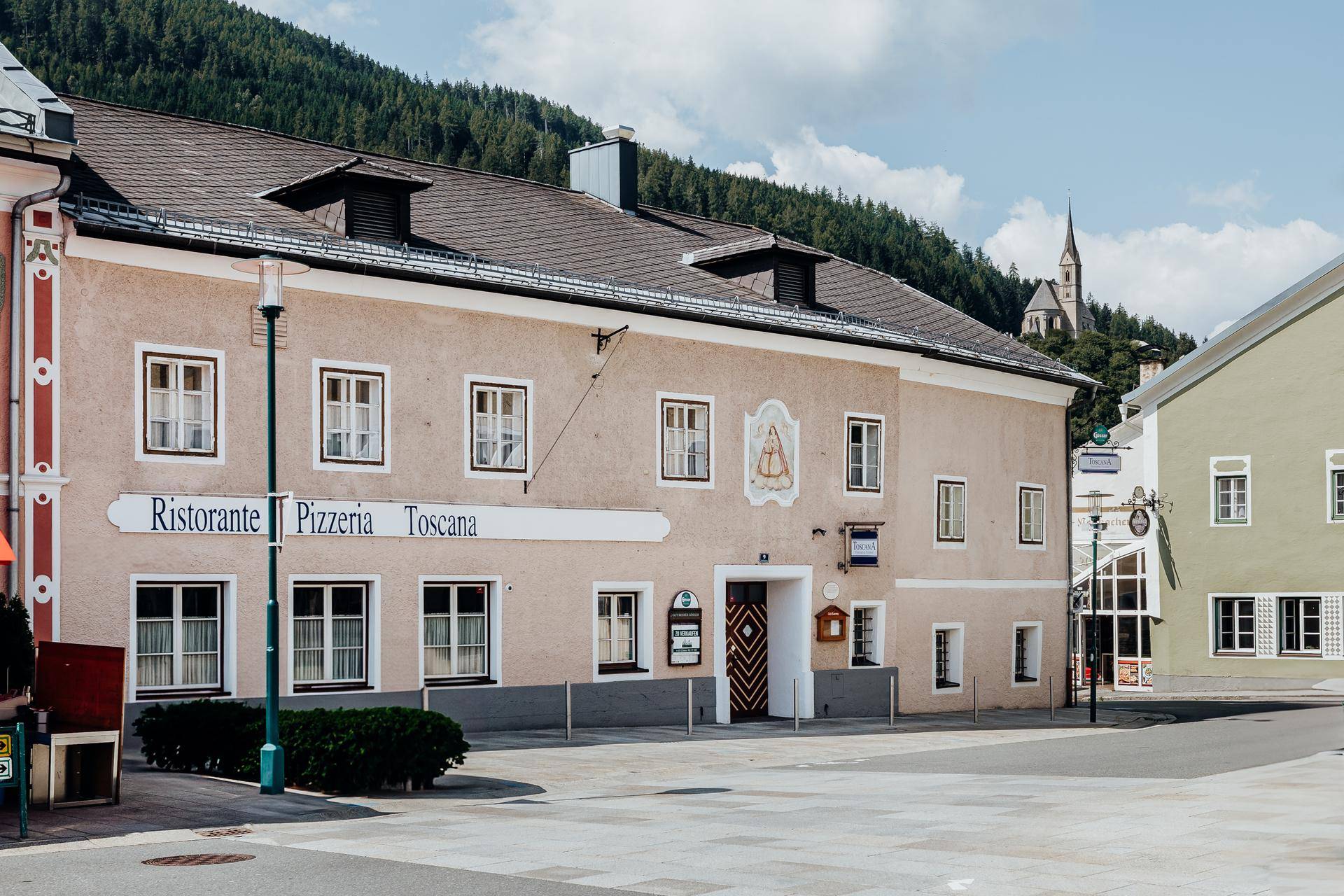 Ansicht des historischen Gebäudes mit Pizzeria-Beschriftung und Bergkulisse im Hintergrund.